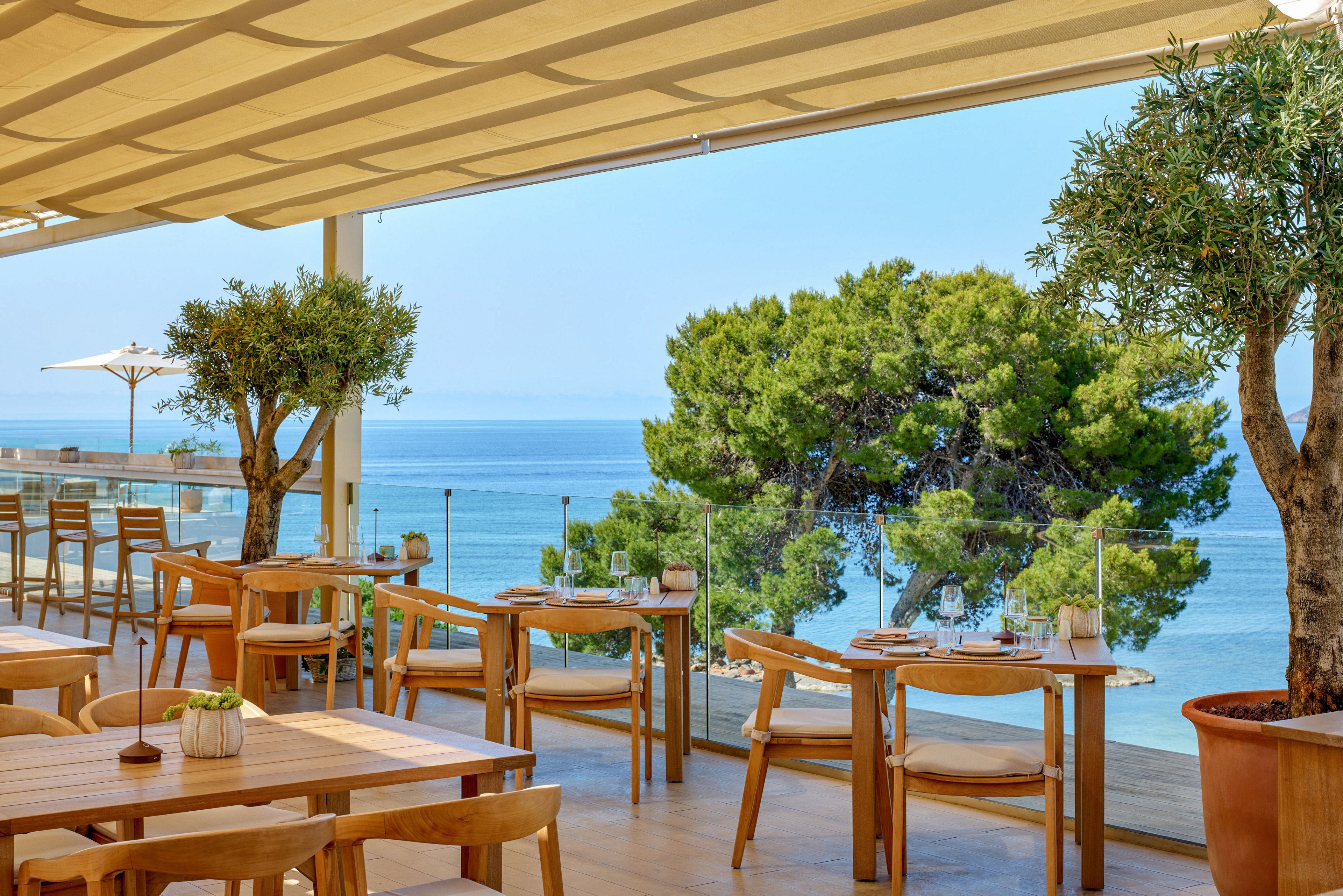 An open-air terrace restaurant overlooks a calm blue sea and lush green trees. Wooden tables and chairs are neatly arranged under a large canopy, creating a shaded dining area. Potted plants and small table decorations add a natural, relaxed touch to the setting. The bright daylight and clear sky suggest a warm, inviting coastal atmosphere ideal for outdoor dining.