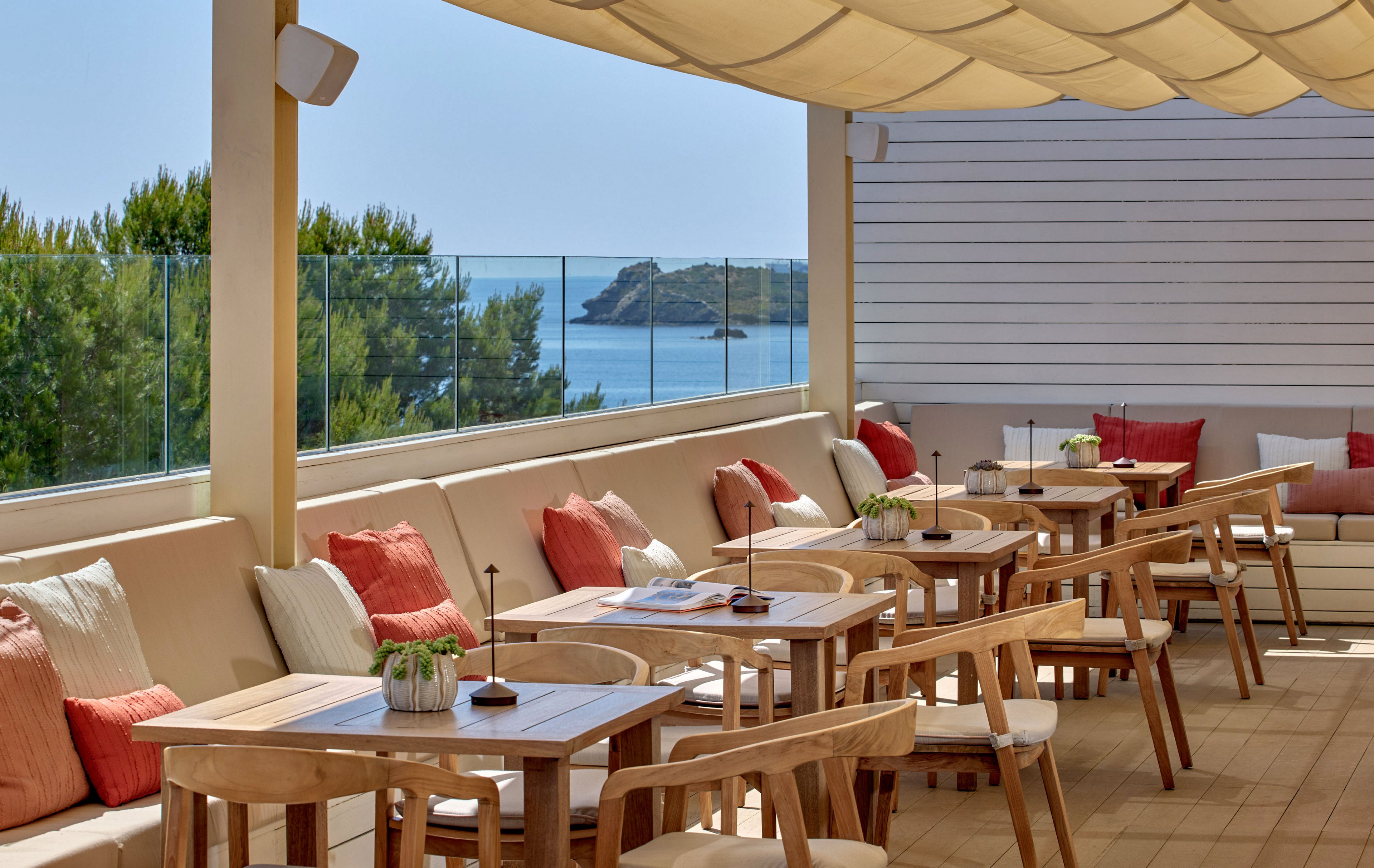 Outdoor restaurant terrace features wooden tables and cushioned bench seating under a light fabric canopy. Red and beige throw pillows add warm color accents against neutral tones. Glass railing reveals a scenic view of the sea, rocky coastline, and green trees in the background. The setting appears calm and inviting, ideal for relaxed al fresco dining.