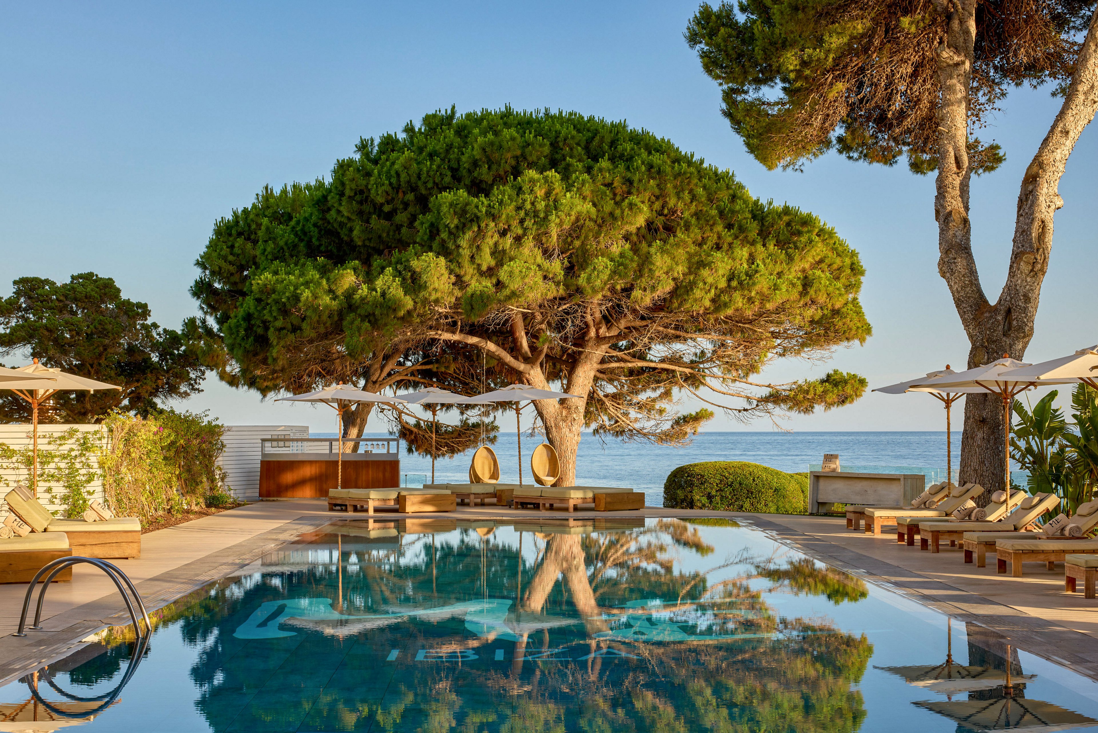 A tranquil outdoor swimming pool reflects a large, lush tree and the clear blue sky. Sun loungers and parasols line the wooden deck, creating a relaxed resort atmosphere beside the water. In the background, the calm sea stretches to the horizon, enhancing the coastal vacation setting. Warm golden light suggests late afternoon or sunset, adding a serene and inviting mood.