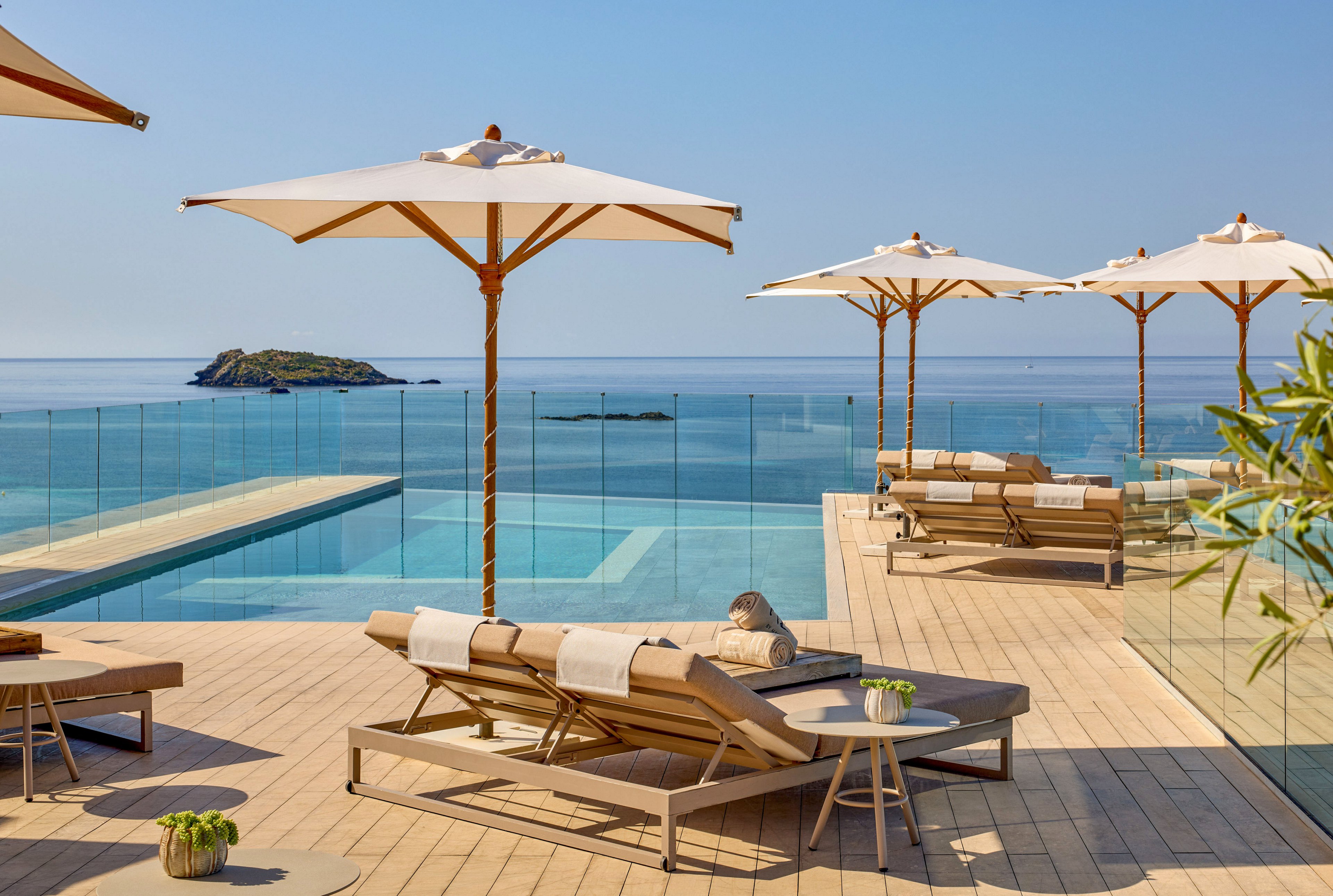 A stylish outdoor pool deck overlooks a calm, blue sea. Wooden sun loungers with cushions are arranged beneath large white umbrellas, facing the water. A glass railing separates the infinity-style pool from the ocean view, with a small island visible in the distance. Potted plants add a touch of greenery to the serene, minimalist setting under clear skies.