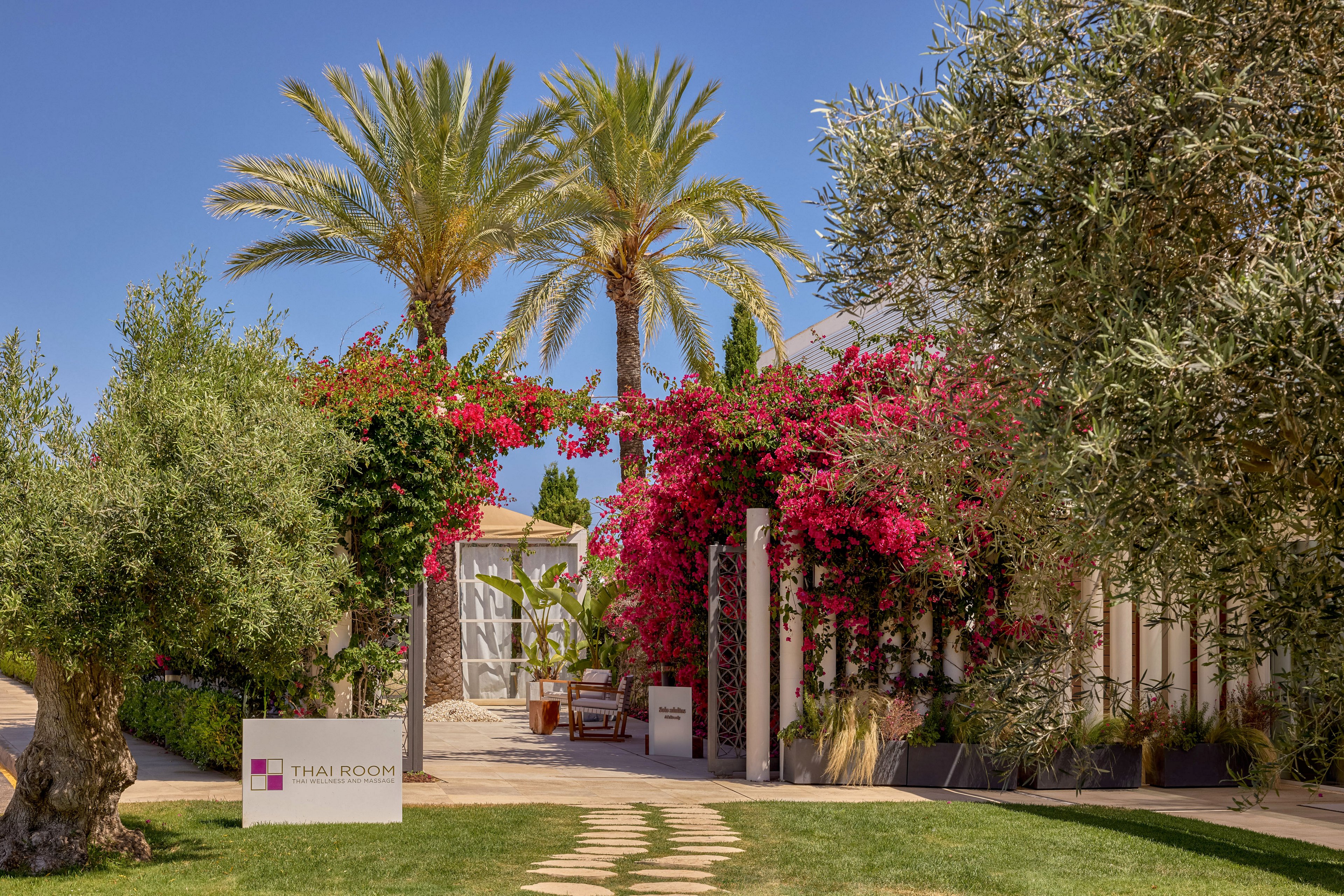 A lush garden pathway leads to an inviting spa entrance labeled "THAI ROOM" on a white sign. Tall palm trees and flowering bougainvillea frame the doorway, creating a vibrant and tropical atmosphere. Outdoor seating is visible inside the entrance, suggesting a relaxing retreat. The scene is bathed in warm sunlight under a clear blue sky, emphasizing a serene vacation setting.