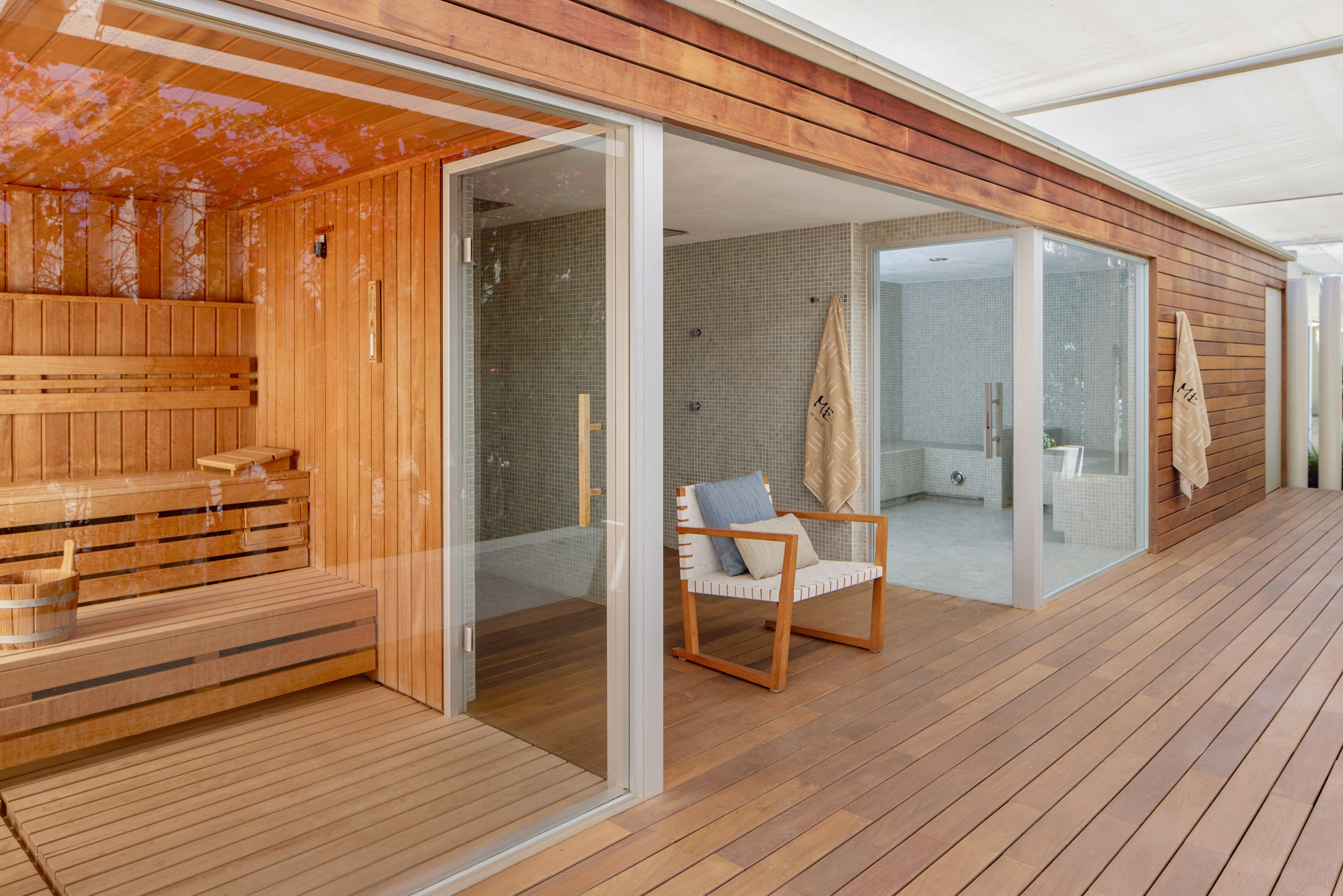 A contemporary spa area features a wooden sauna with glass doors and built-in bench seating. Adjacent to the sauna is a minimalist relaxation space with a cushioned armchair and light-toned walls. Large floor-to-ceiling windows open onto a spacious wooden deck, creating an airy, indoor-outdoor feel. The clean lines, natural wood, and neutral colors convey a calm, luxurious atmosphere ideal for wellness and relaxation.
