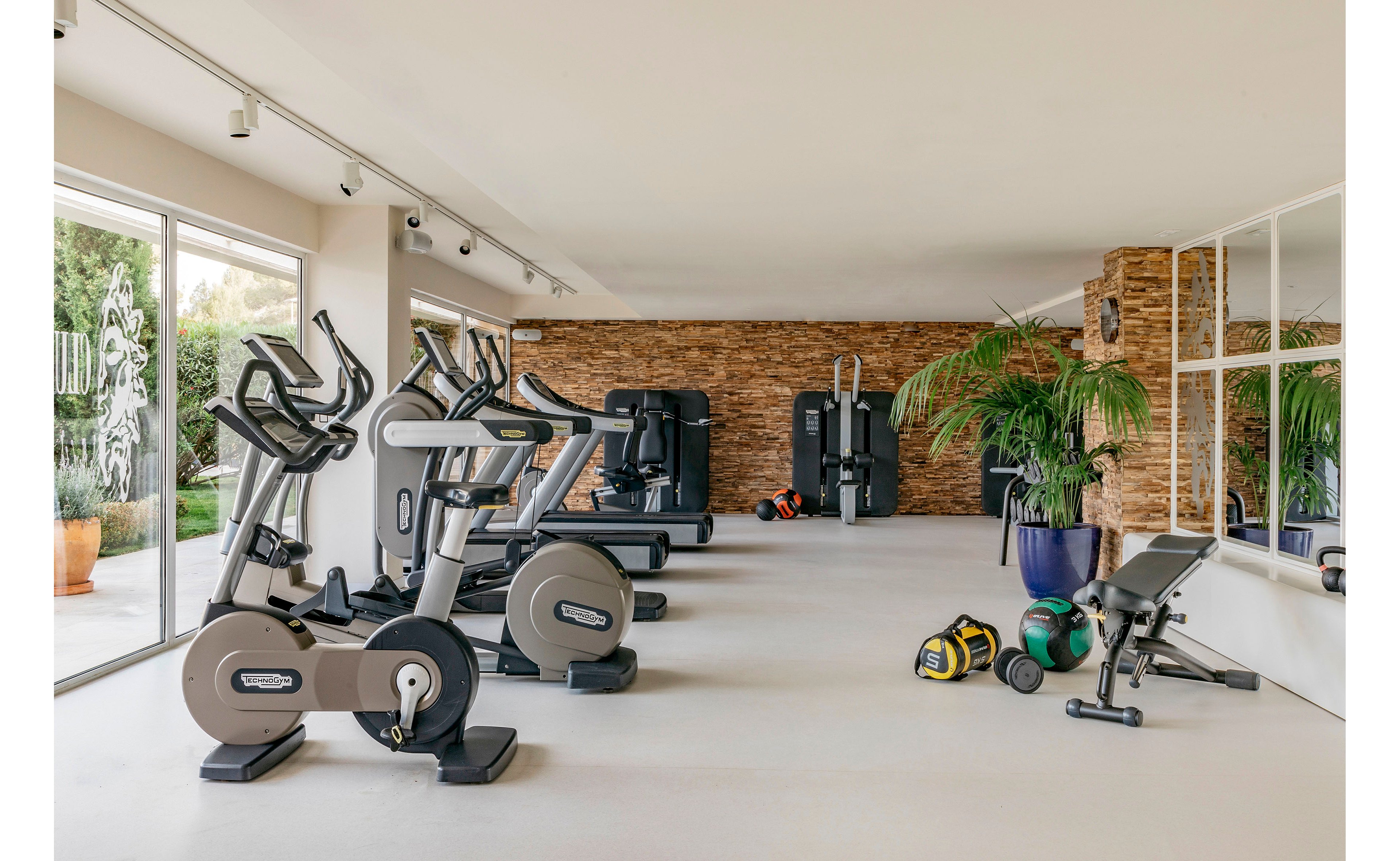 Bright, spacious fitness room featuring multiple exercise bikes and elliptical machines along one side. Strength training equipment, kettlebells, medicine balls, and a weight bench are arranged near a stone accent wall. Large floor-to-ceiling windows let in natural light and reveal greenery outside. Indoor plants and clean, minimal decor create a contemporary, inviting workout environment.