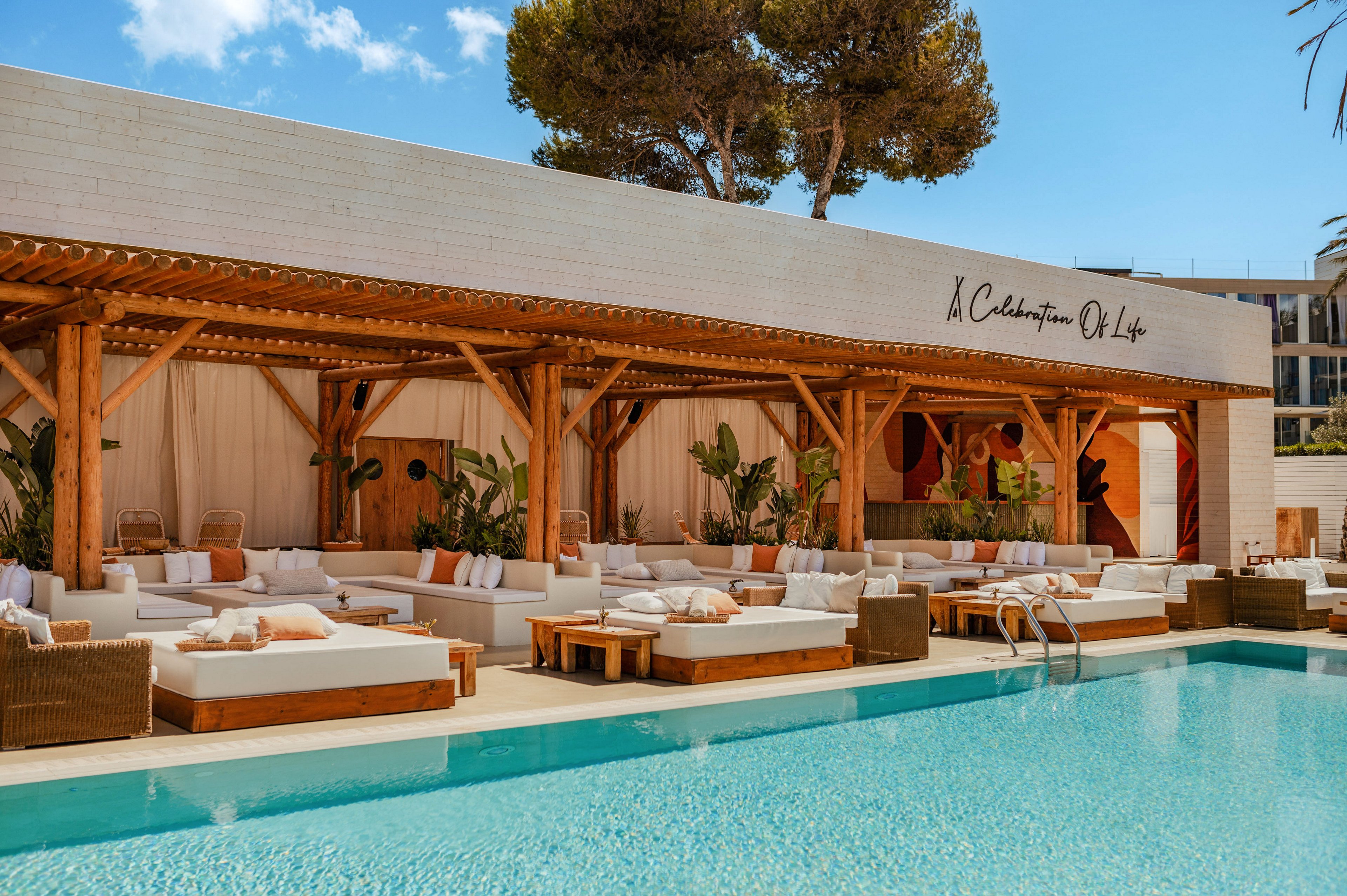 An upscale outdoor lounge area features white cushioned daybeds and wooden cabanas beside a clear blue swimming pool. The setting appears to be a stylish resort or beach club with natural wood structures and tropical plants. Neutral-toned pillows and draped curtains create a relaxed, elegant atmosphere. A handwritten-style sign is visible on the building facade, adding to the curated, contemporary design.