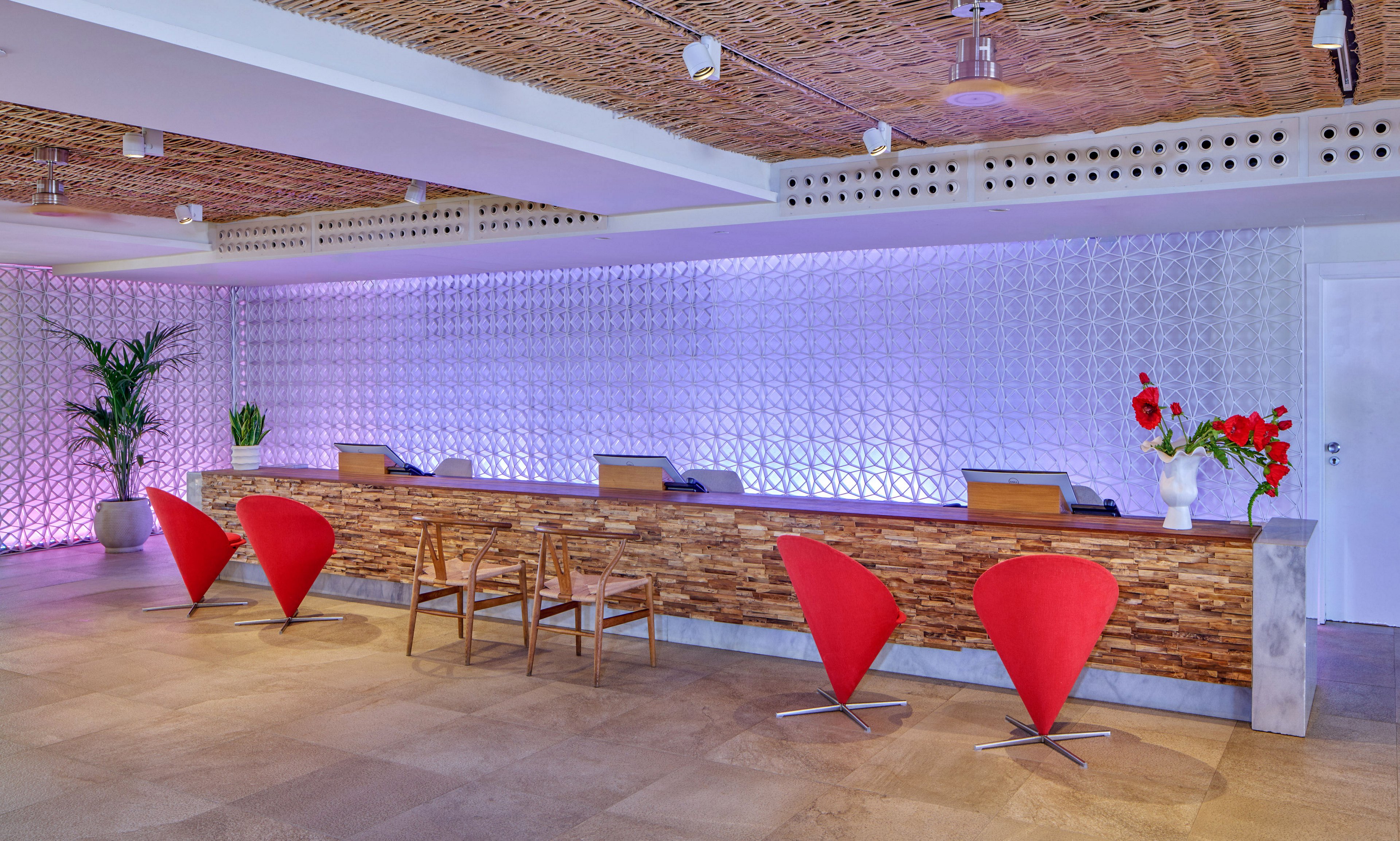 Contemporary reception desk area featuring a long wooden counter with multiple workstations. The space is highlighted by sculptural red chairs and simple wooden chairs in front of the desk. A textured white wall with soft purple lighting creates a stylish backdrop, while a potted plant and red flowers add natural touches. The ceiling and floor use warm, earthy materials, giving the lobby a welcoming, modern atmosphere.