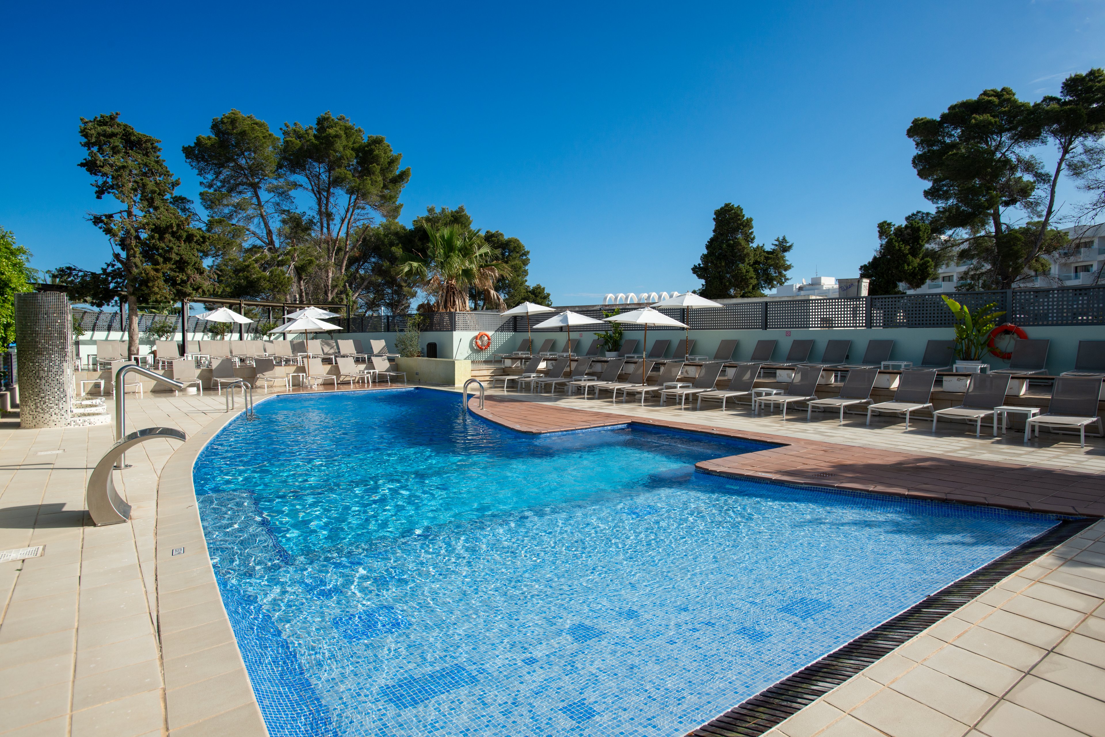 An outdoor swimming pool glistens under a clear blue sky at a modern hotel or resort. Numerous sun loungers and white umbrellas line the tiled pool deck, suggesting a relaxing vacation setting. Tall trees and a light-colored privacy wall frame the area, creating a secluded atmosphere. The calm water and empty chairs evoke a peaceful, early-morning or off-peak moment by the pool.