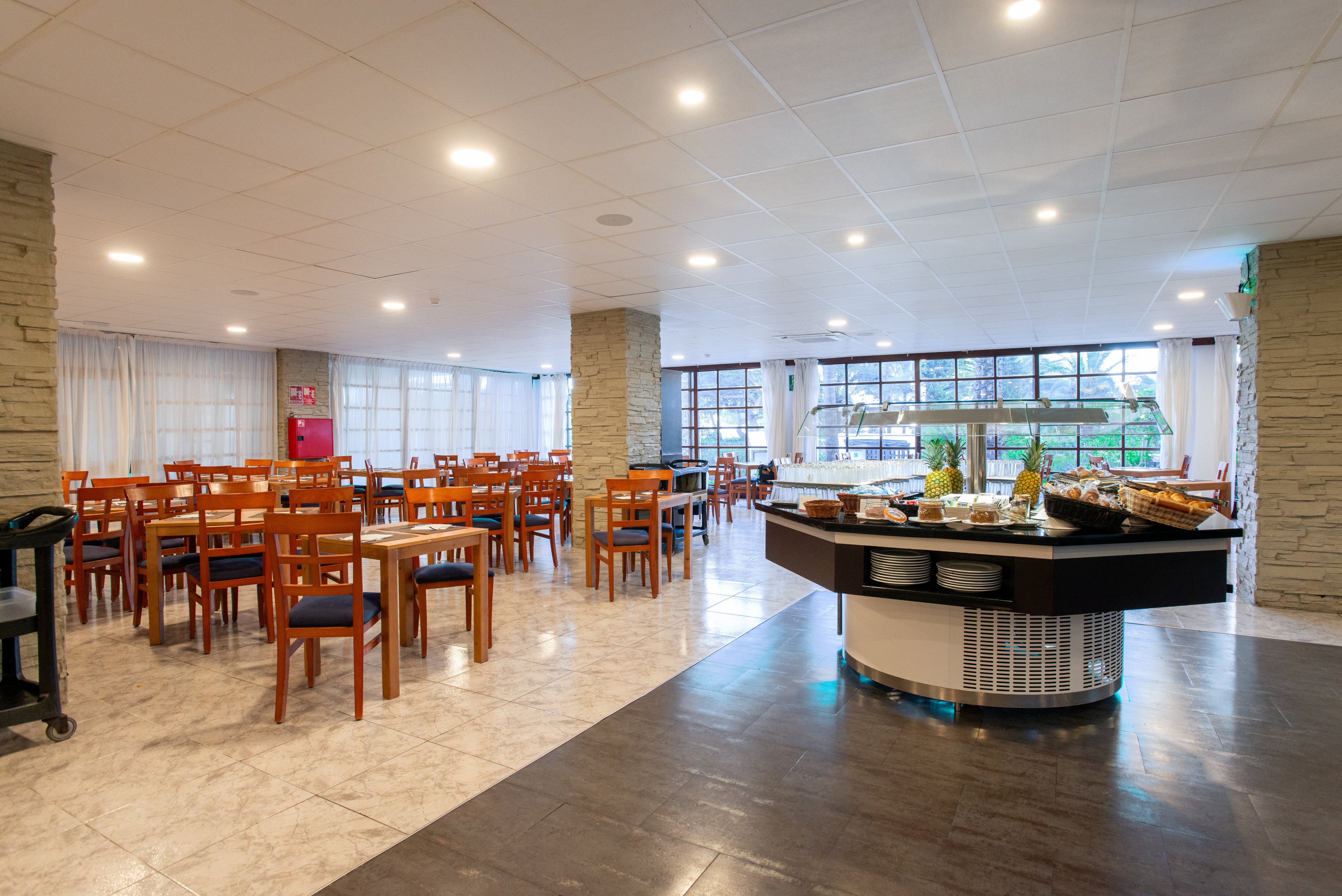 Spacious, modern restaurant dining area with wooden tables and chairs stands empty, ready for guests. A central circular buffet island displays breakfast items, plates, and serving dishes. Large windows along the back wall let in natural light and reveal greenery outside. Neutral tones, stone columns, and a clean tiled floor create a calm, welcoming atmosphere.