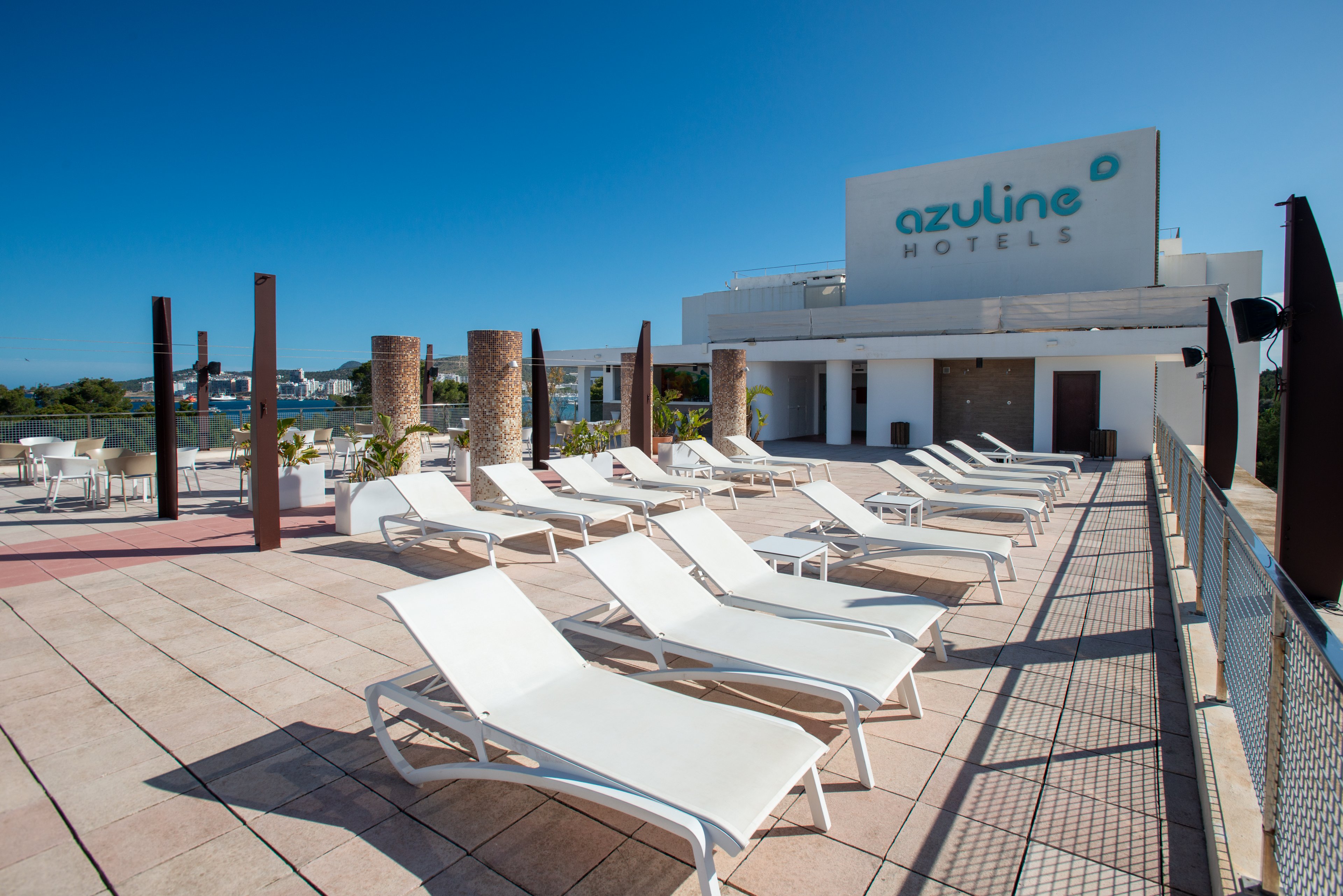 A spacious rooftop terrace at Azuline Hotels features multiple white sun loungers arranged in rows under a clear blue sky. The area is paved with light tiles and bordered by a safety railing overlooking the surrounding town and water. Vertical wooden posts and stone columns add architectural interest to the open-air deck. The large Azuline Hotels sign is prominently visible on the building facade in the background.