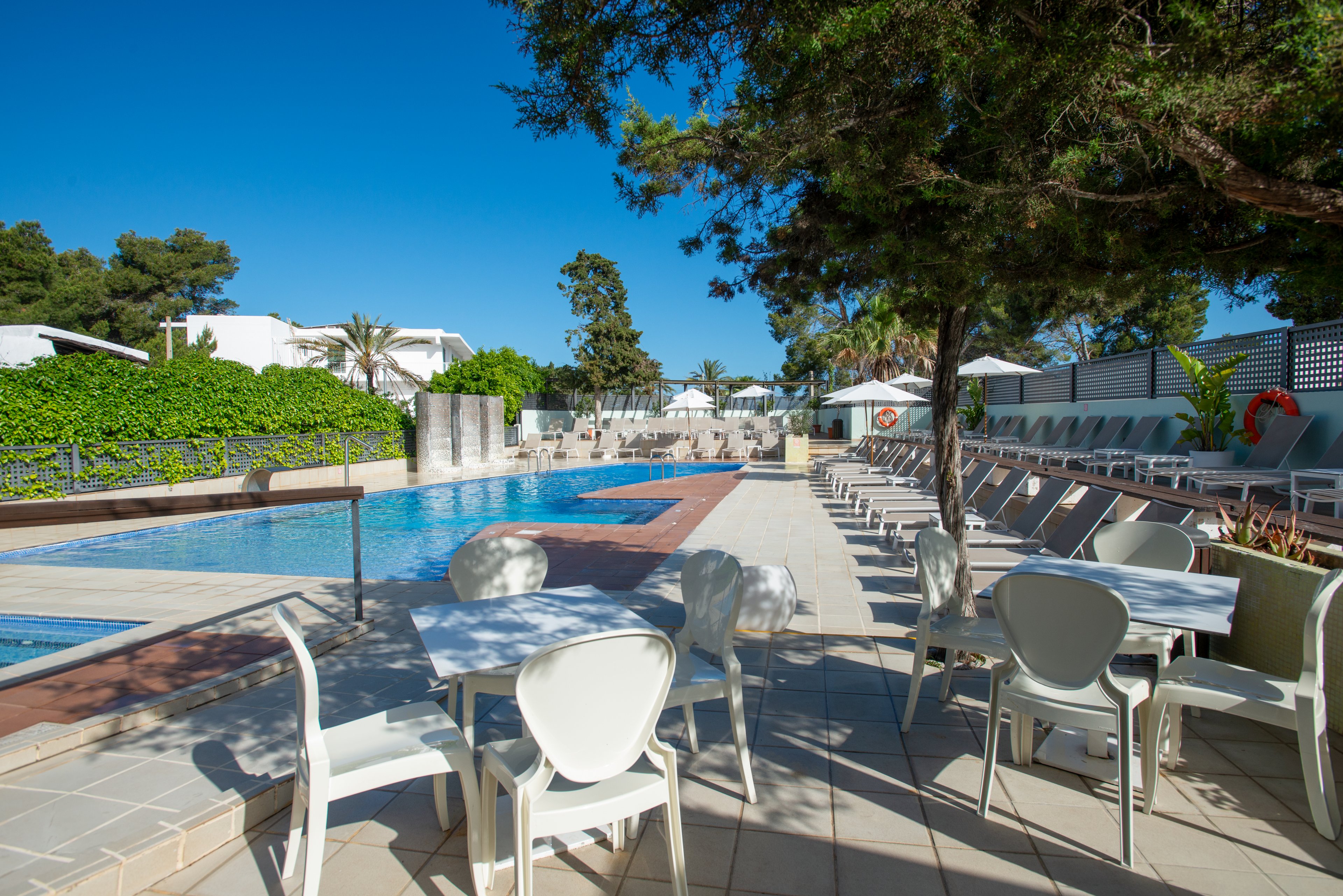 Outdoor resort swimming pool area on a clear sunny day. White lounge chairs line the pool deck beside a row of green hedges and modern low-rise buildings. A shaded seating area with white tables and chairs sits in the foreground under tall trees. The scene feels calm and inviting, ideal for relaxation and vacation leisure.