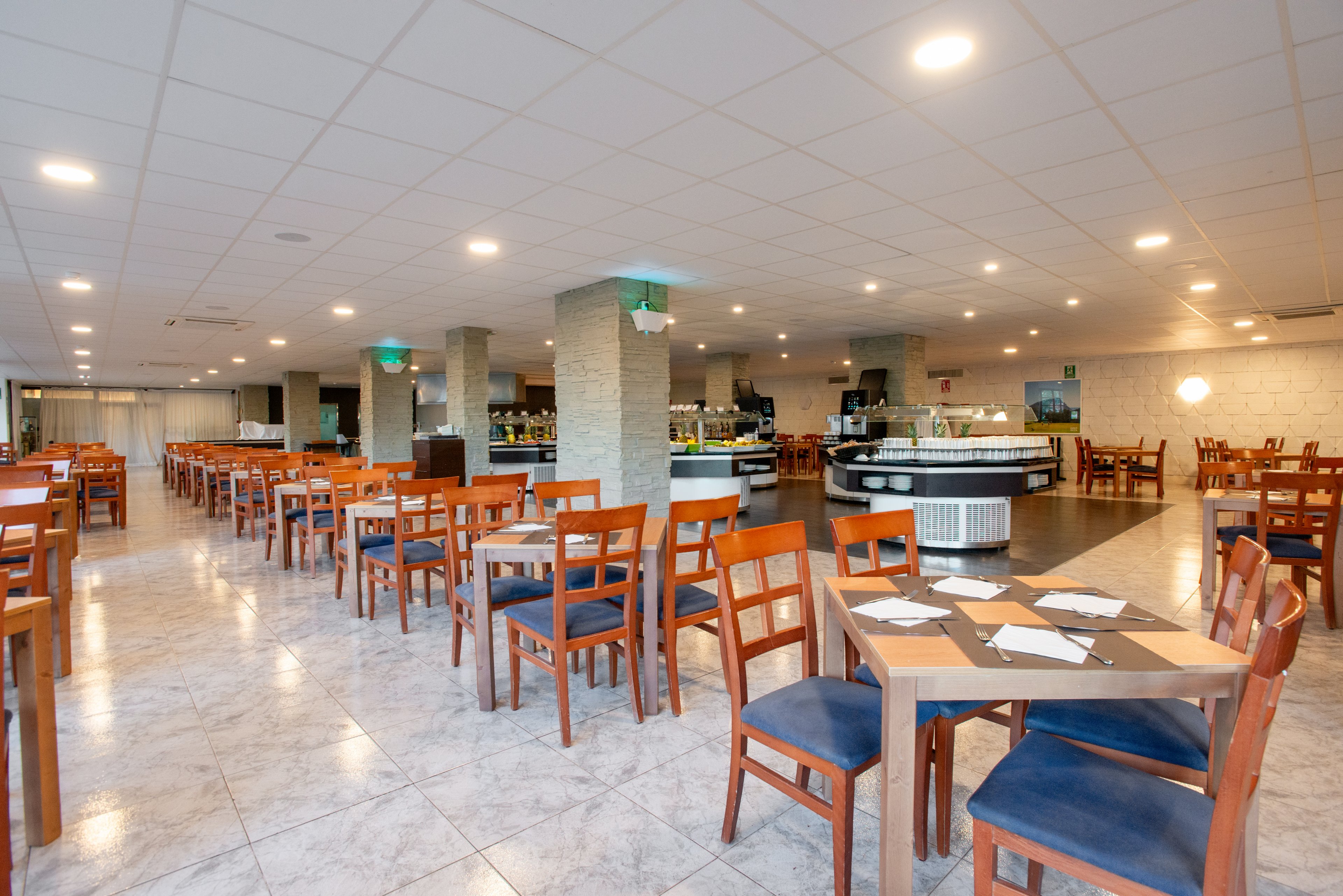 Wide view of an empty restaurant dining room with neatly arranged wooden tables and chairs. The space features a tiled floor, light-colored walls, and a grid ceiling with recessed lighting. A buffet or serving area is visible in the background with counters and displays. The overall atmosphere is clean, orderly, and ready to welcome guests.