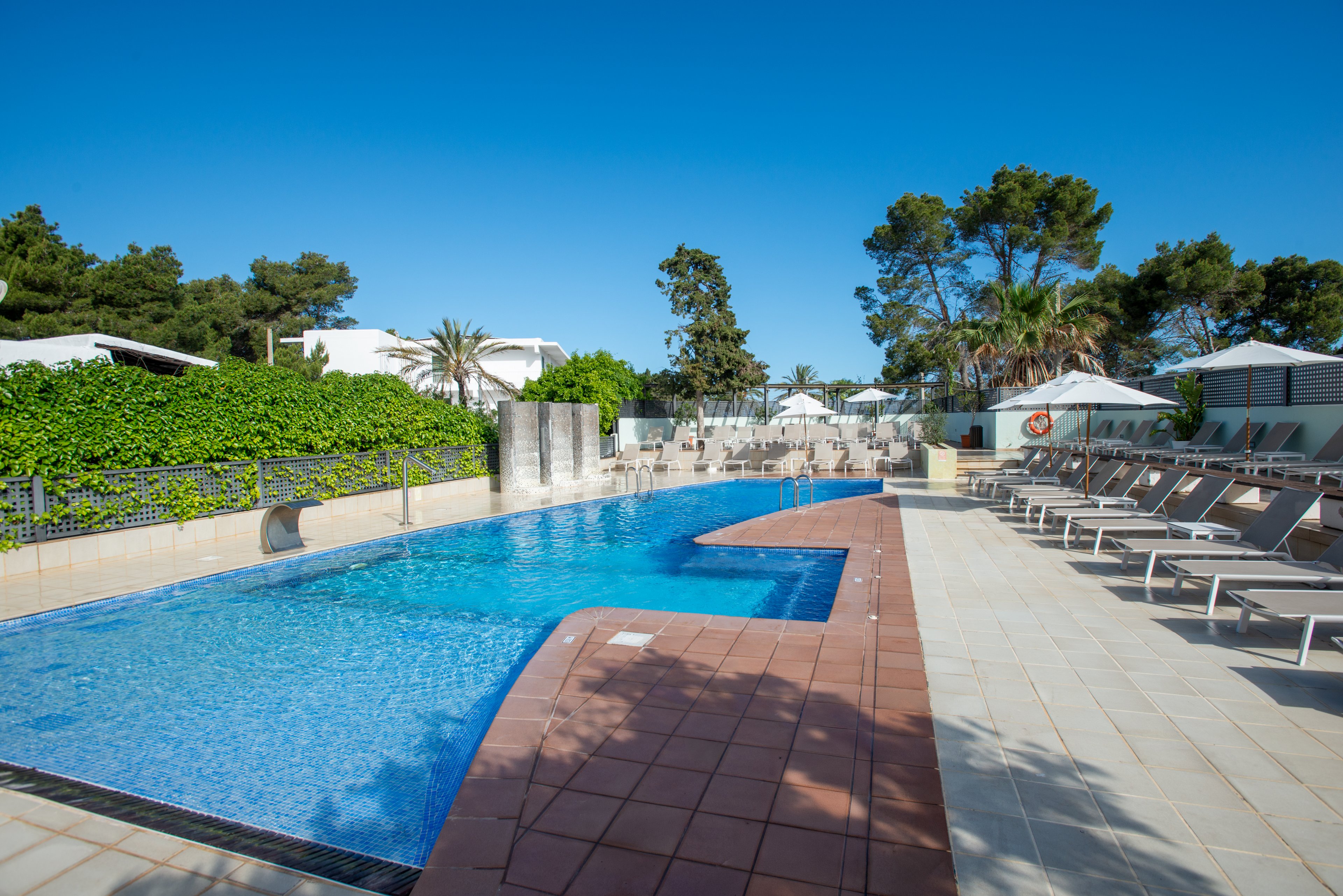 An outdoor swimming pool stretches alongside a row of empty lounge chairs at a resort. The clear blue water reflects the bright sky, framed by tiled poolside decking and safety railings. White umbrellas and low white buildings sit behind a lush green hedge, with tall trees in the background. The scene suggests a calm, sunny day ideal for relaxing by the pool.