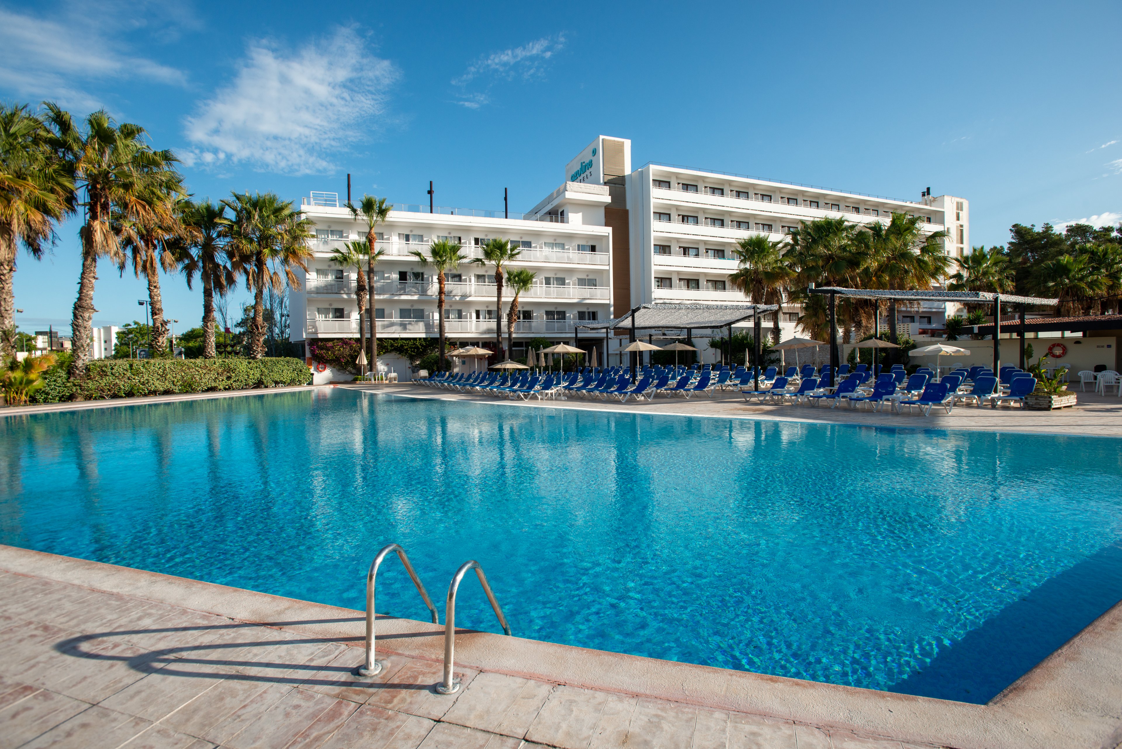 A large outdoor swimming pool dominates the foreground of a modern resort hotel. Rows of blue lounge chairs and umbrellas line the poolside deck, suggesting a relaxing vacation setting. Tall palm trees frame the scene, adding a tropical atmosphere under a clear blue sky. The multi-story hotel building in the background features balconies and wide windows overlooking the pool area.
