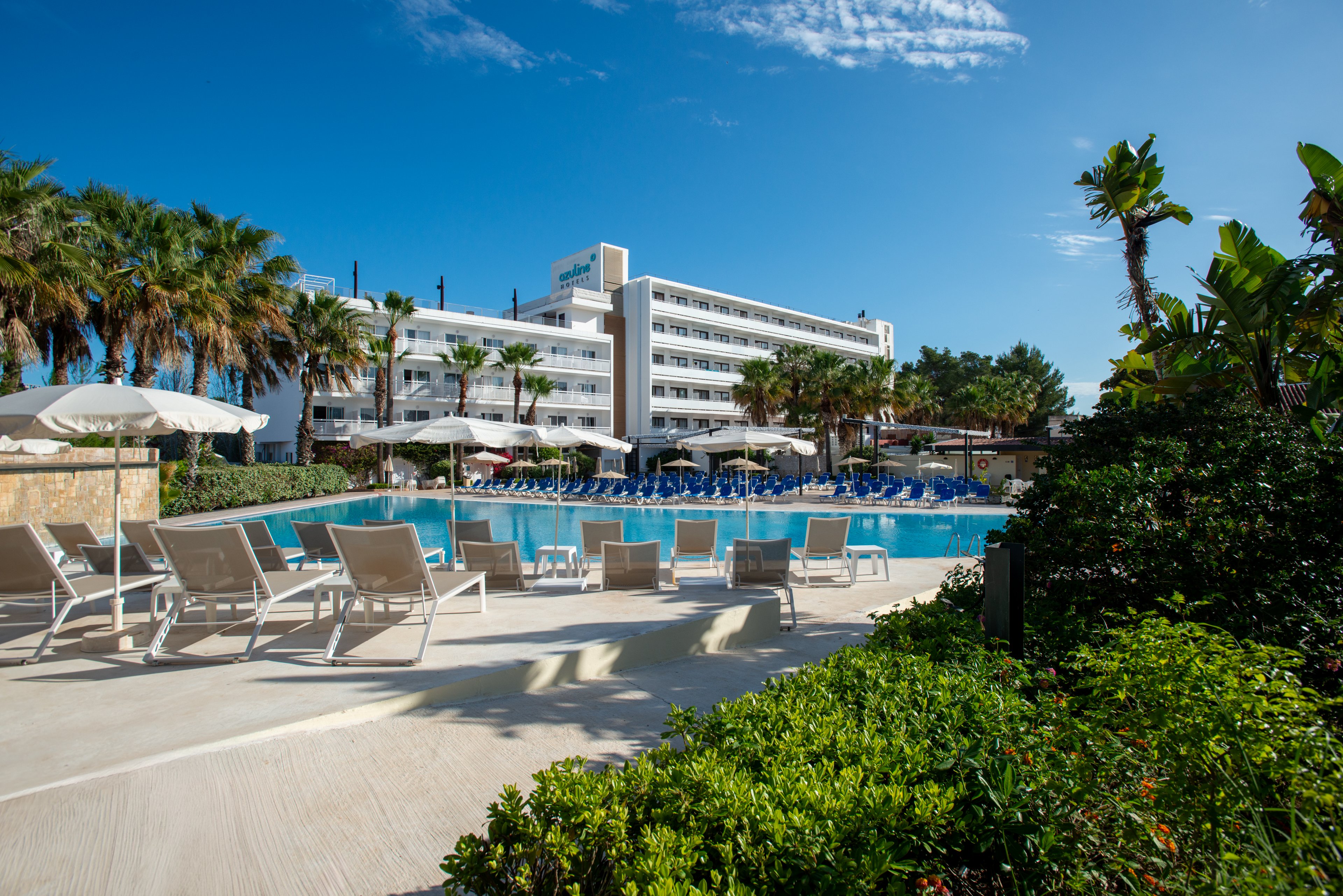 A contemporary multi-story resort hotel overlooks a large outdoor swimming pool. White sun loungers and umbrellas line the poolside terrace, creating a relaxing vacation setting. Lush green plants and palm trees frame the scene under a bright blue sky with scattered clouds. The atmosphere suggests a sunny, tranquil holiday destination ideal for leisure and relaxation.