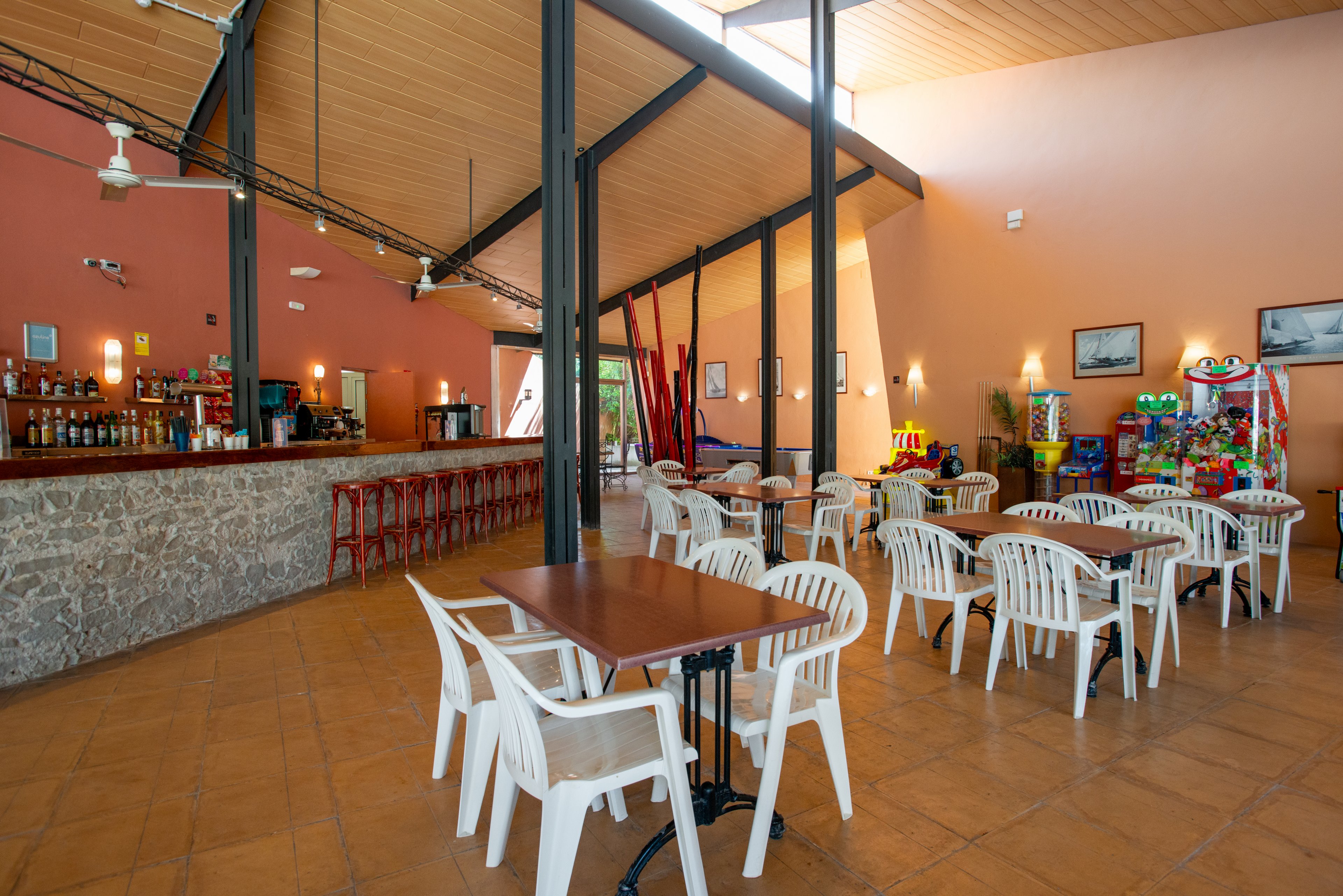 Bright, open bar area with a long stone counter and rows of red barstools. White plastic tables and chairs are arranged across the tiled floor, creating a casual dining space. The high wooden ceiling and exposed beams give the room an airy, modern feel. In the background, colorful arcade machines and decor add a playful touch to the relaxed atmosphere.