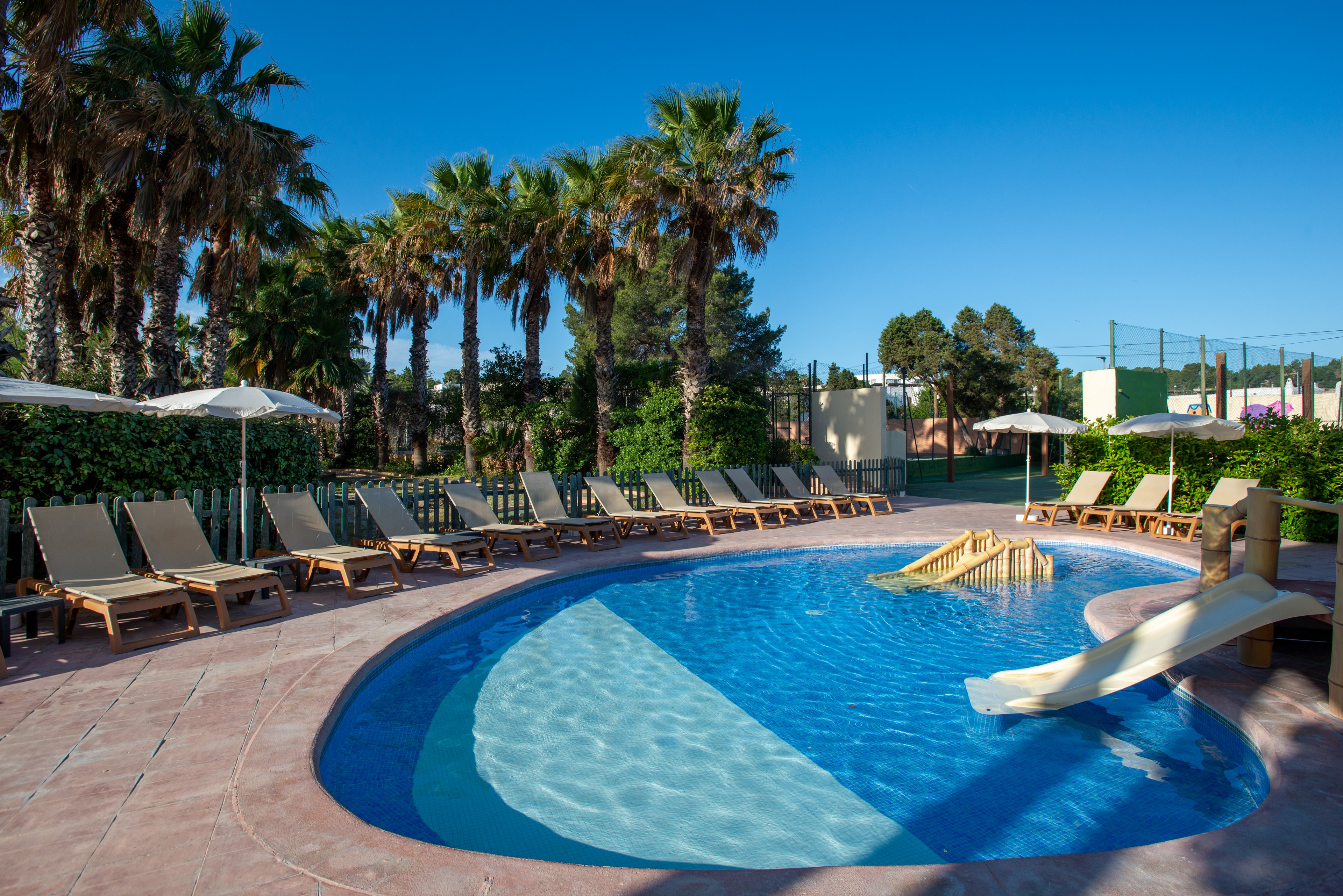 A small circular outdoor swimming pool with a gentle slide is surrounded by numerous reclining lounge chairs. White umbrellas provide shade along the poolside, while tall palm trees line the background. The clear blue water reflects the bright sky, creating a relaxing vacation atmosphere. Green hedges and distant sports courts suggest a larger resort or hotel complex.