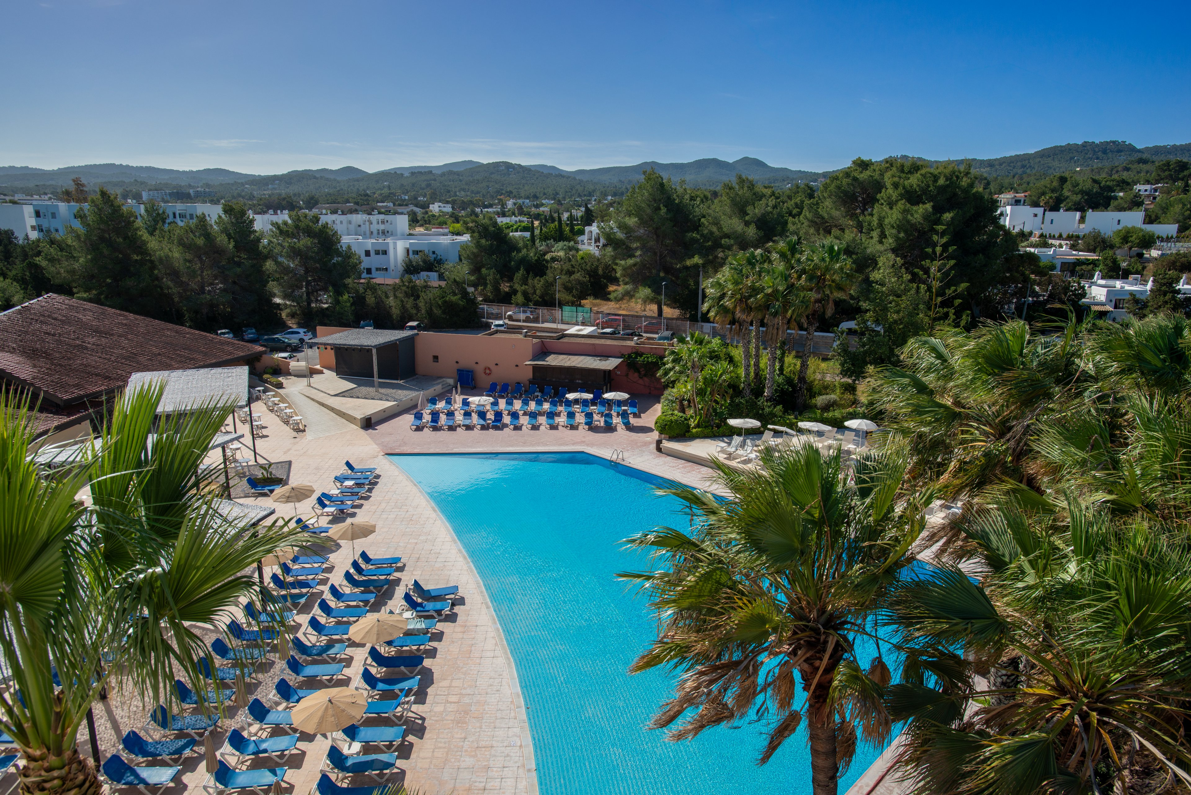A large outdoor swimming pool is surrounded by rows of blue sun loungers and umbrellas at a resort. Palm trees frame the pool area, adding a tropical feel to the scene. In the background, low white buildings sit among dense green trees and rolling hills. The clear blue sky and bright sunlight suggest a warm, relaxing vacation atmosphere.