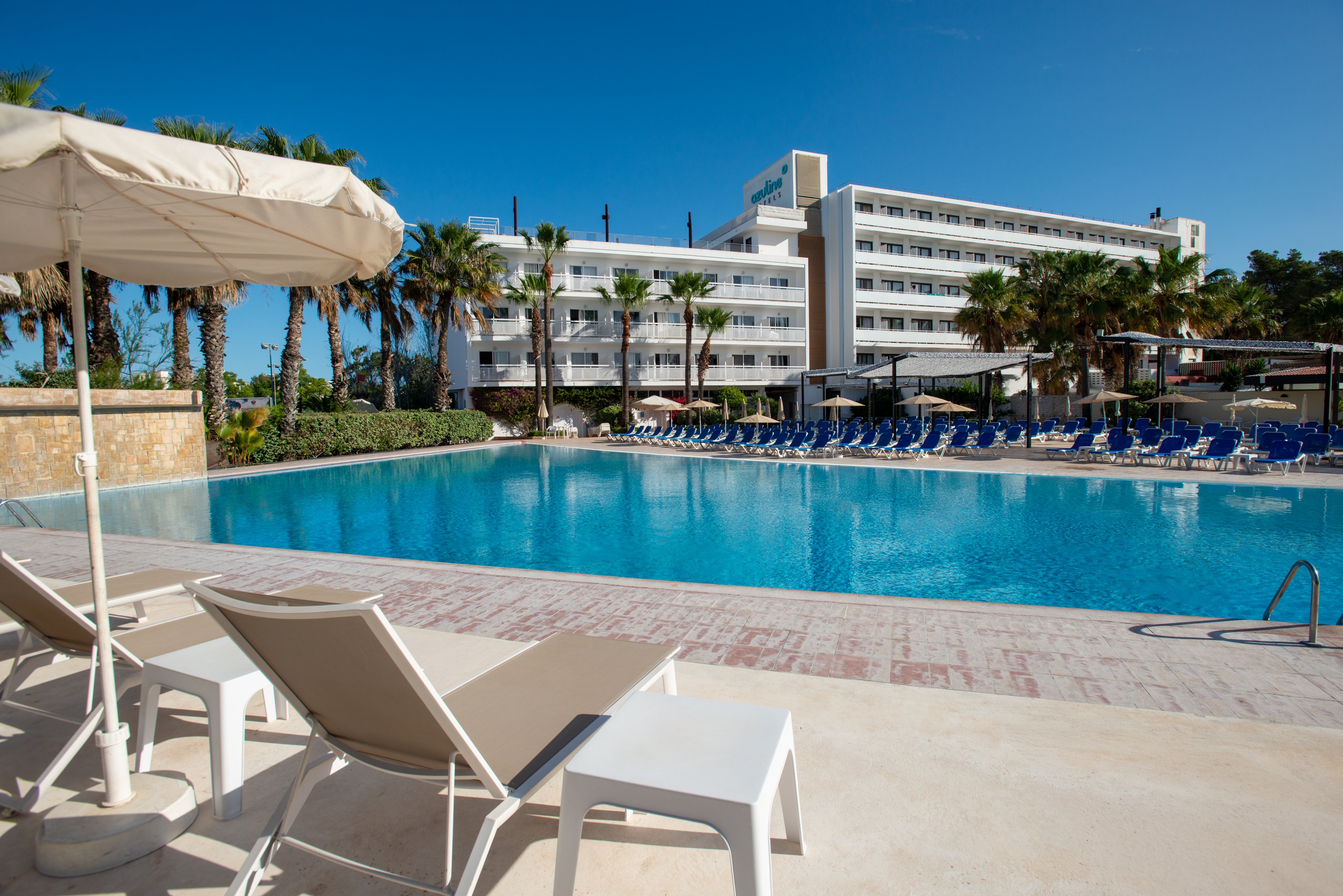 A spacious outdoor swimming pool sits in front of a multi-story resort hotel under a clear blue sky. White lounge chairs and umbrellas line the pool deck, with rows of blue sunbeds visible across the water. Tall palm trees frame the scene, adding a tropical atmosphere to the modern property. The bright sunlight and calm water create a relaxing vacation setting.