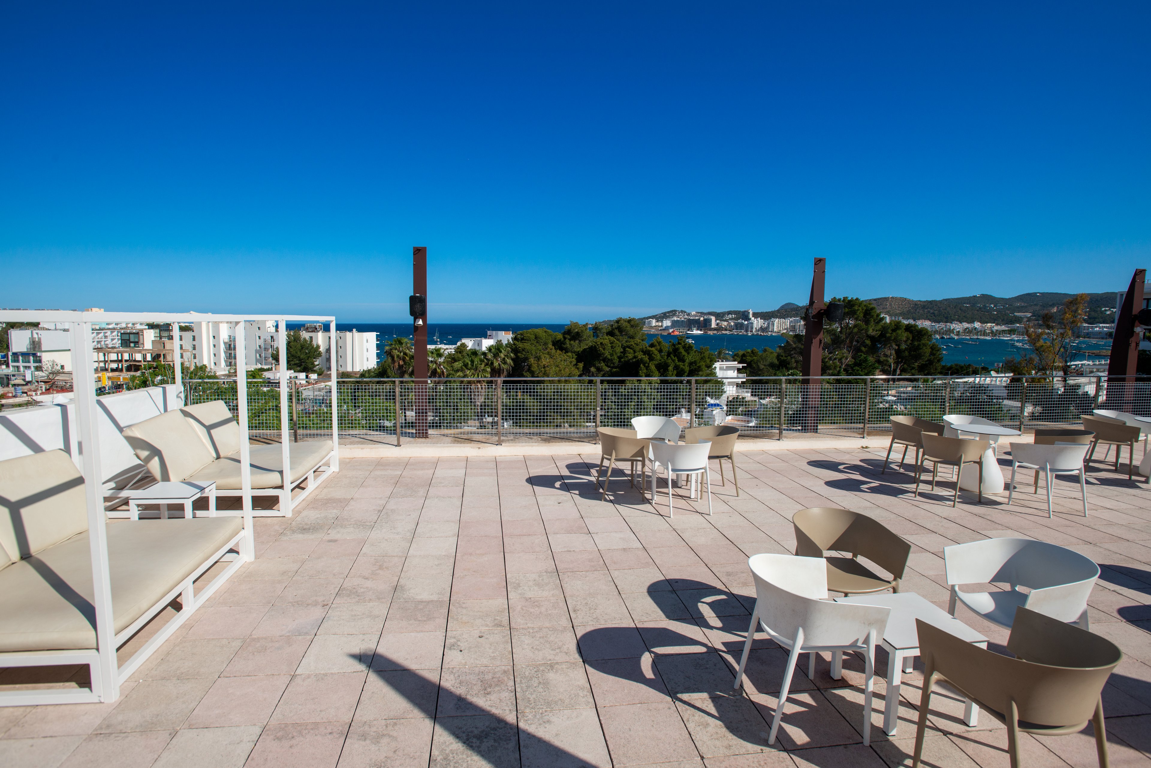 An open rooftop terrace features modern white lounge beds and round tables with chairs. The space is paved with light stone tiles and bordered by a glass railing. In the distance, a coastal cityscape and blue sea are visible under a clear sky. The scene suggests a relaxing outdoor leisure area with panoramic ocean views.