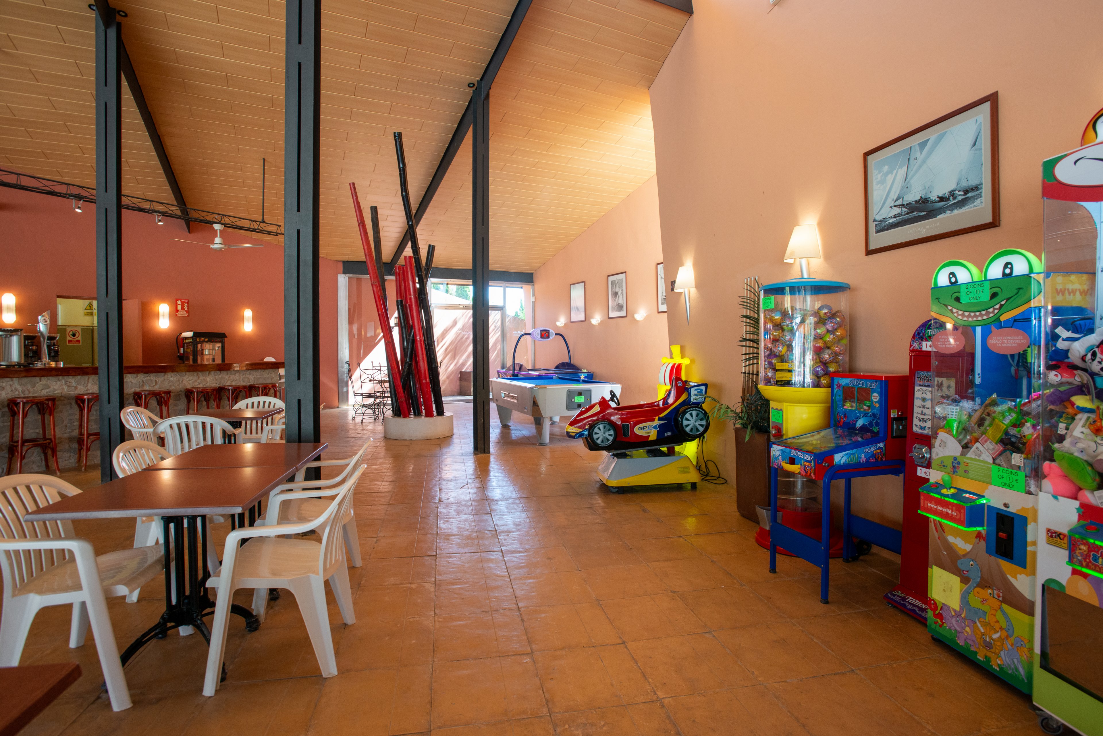 Bright indoor recreation room featuring several arcade and vending machines along one wall. A small children's ride-on car and a foosball or air hockey table sit near the center of the space. On the left, plastic chairs and tables create a casual seating area beside a bar counter. Warm peach walls, tiled floor, and nautical artwork contribute to a relaxed holiday atmosphere. The room appears empty, ready for families or guests to enjoy games and refreshments.