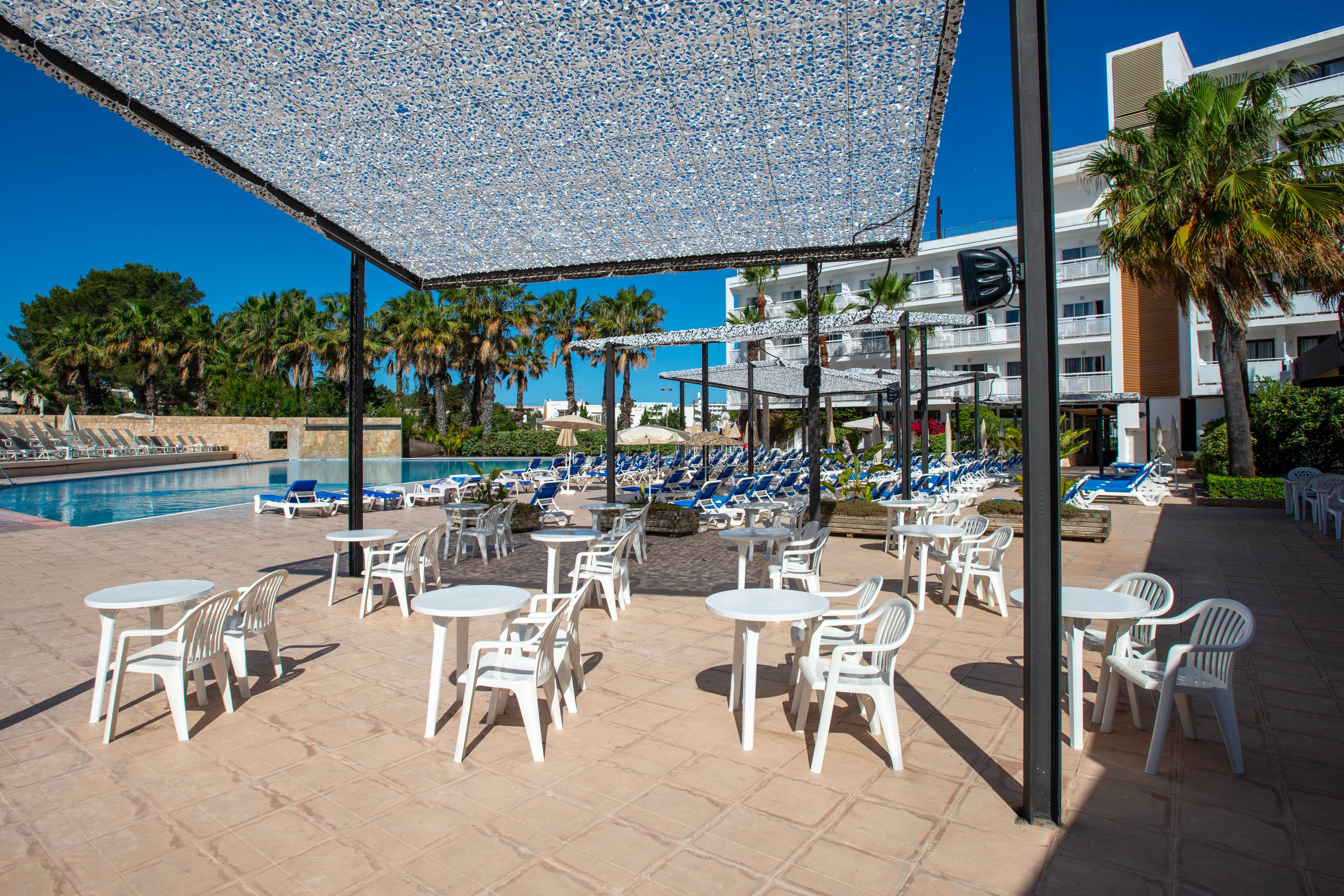 An outdoor resort terrace features white plastic tables and chairs arranged beneath a modern shade canopy. In the background, rows of blue sun loungers line a large swimming pool. Palm trees and landscaped greenery surround the area, adding a tropical feel. A multi-story hotel building with balconies overlooks the bright, sunlit pool deck under a clear blue sky.
