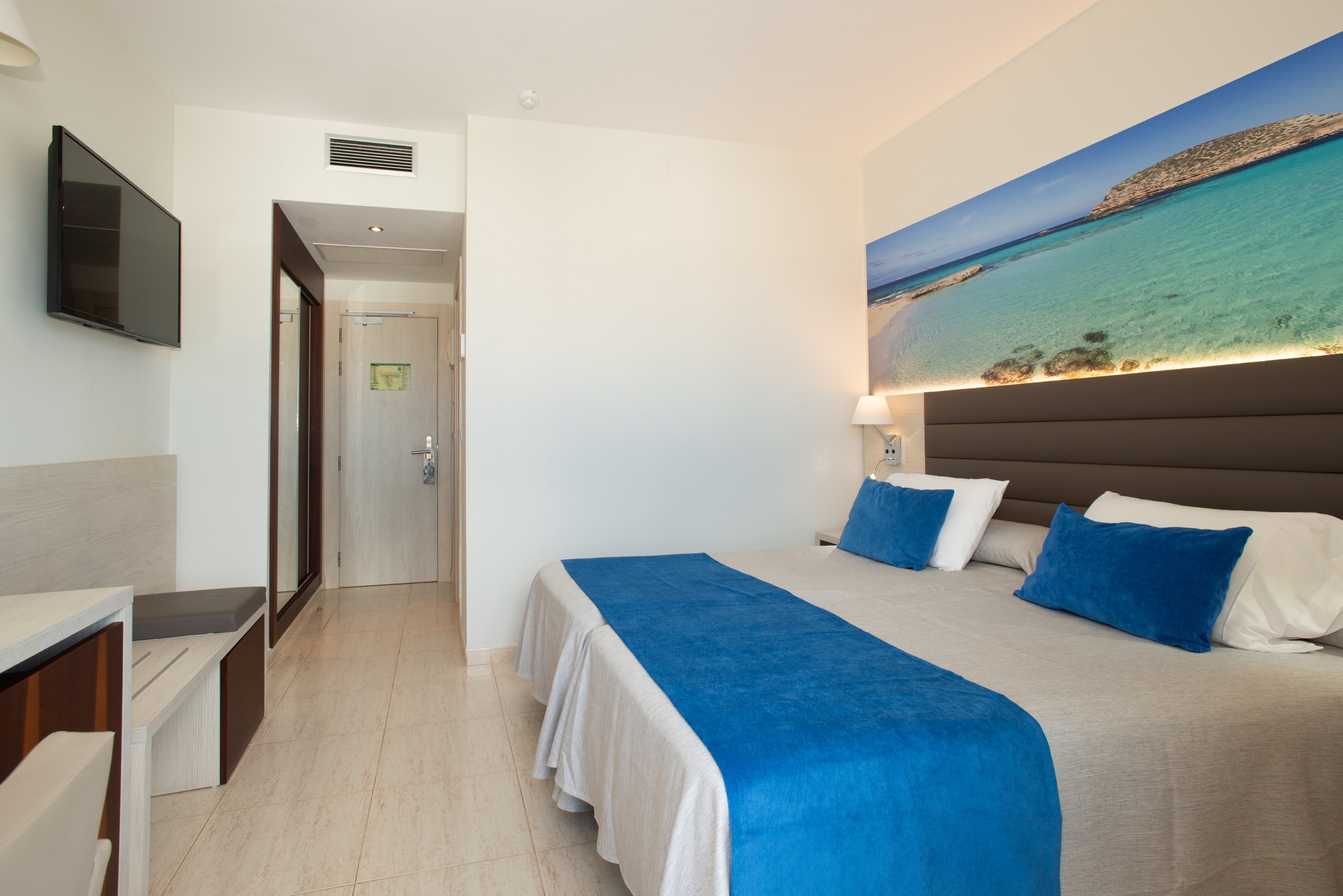 A clean, modern hotel room features two twin beds pushed together with white linens and bright blue throws and pillows. The space is decorated in neutral tones with a large wall art print of a tropical beach above the headboard. A flat-screen television is mounted on the wall opposite the beds, and a small desk area sits to the side. The room leads to a hallway with a door, creating a sense of depth and inviting entry.