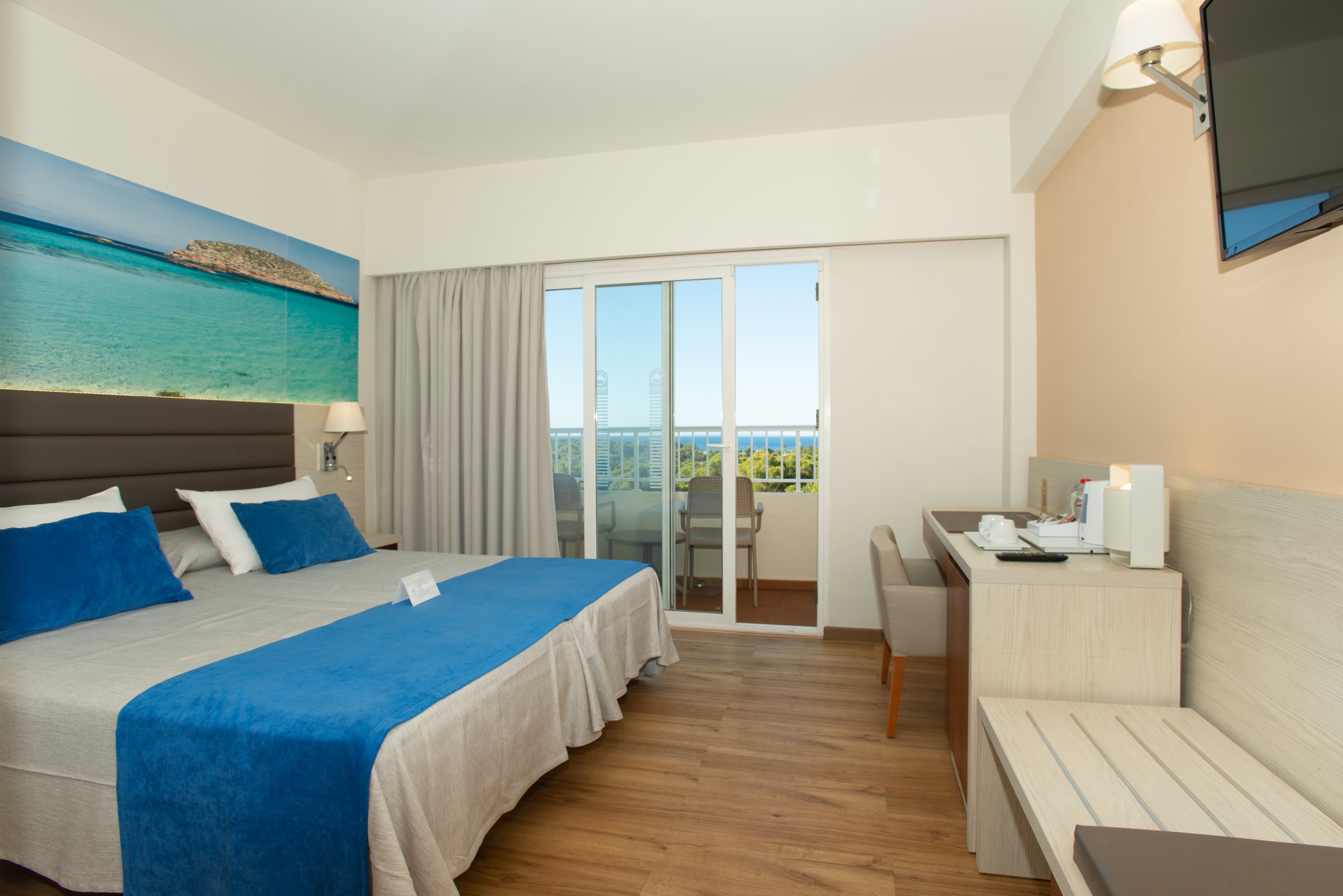 Modern hotel room featuring a double bed with blue and white linens is shown in daylight. A large sliding glass door opens to a balcony with chairs and a distant view of the sea and greenery. The interior includes wooden flooring, a light-colored desk with a kettle and cups, and a wall-mounted television. A wide seascape artwork above the bed and neutral walls create a clean, relaxing atmosphere.