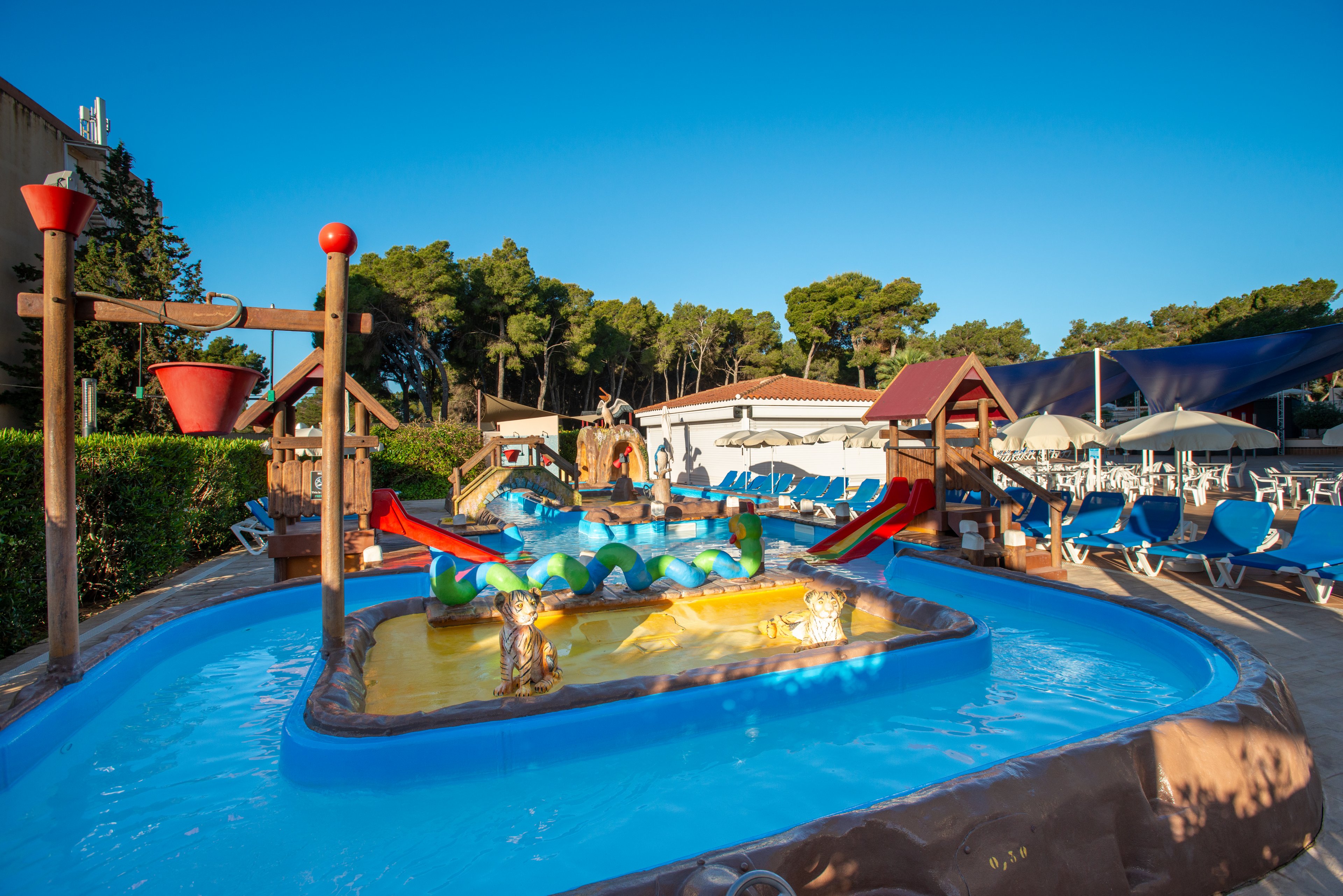 An outdoor children’s splash pool features shallow blue water and colorful play structures. Small slides, water-spraying toys, and tipping buckets create a fun aquatic playground. Rows of blue sun loungers and umbrellas line the background near a building. Tall trees and a clear blue sky suggest a warm, sunny day at a family-friendly resort or campground.