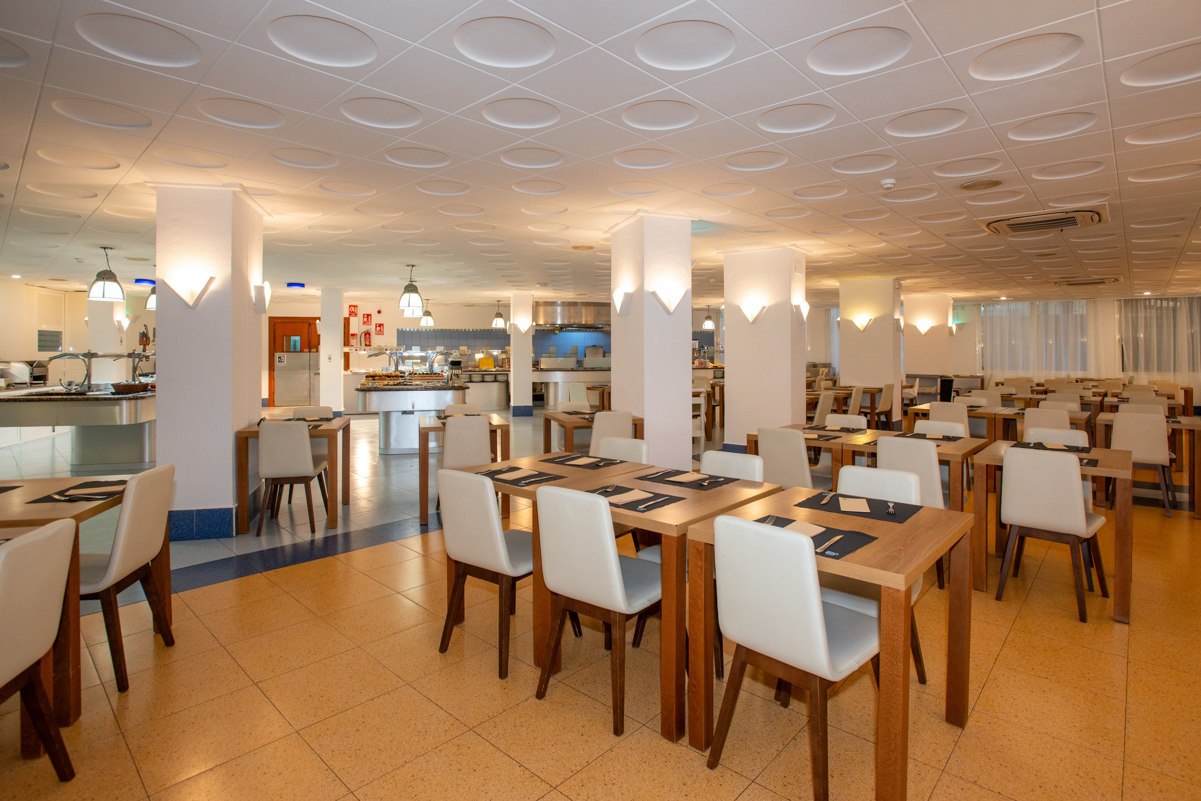 Spacious restaurant dining room with neatly arranged wooden tables and white upholstered chairs. The room is empty of people, emphasizing the clean and orderly setup. Warm ceiling and wall lights create a cozy atmosphere, while the open buffet or serving area is visible in the background. The overall design feels contemporary and inviting, suitable for hotel or cafeteria dining.