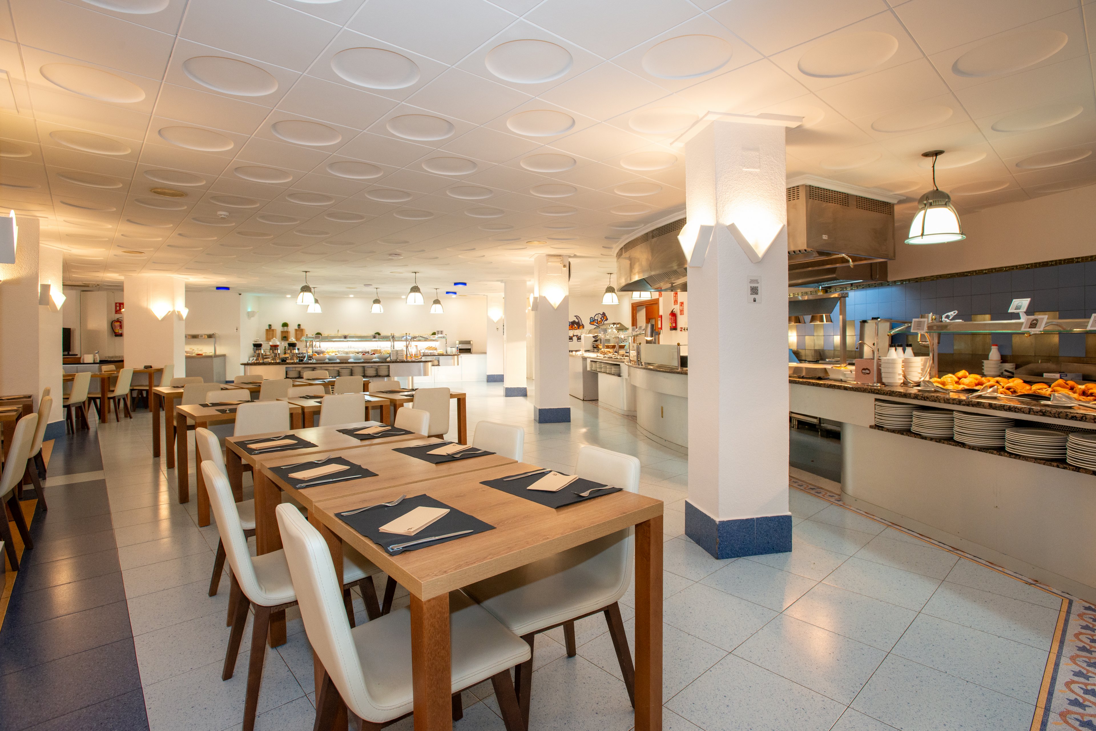 Spacious self-service restaurant interior featuring wooden tables and white chairs. A long buffet counter on the right displays plates and various food trays under warm lighting. The open kitchen area in the background shows staff preparing meals. Clean white walls, tiled floor, and modern ceiling lights create a bright, contemporary dining atmosphere.