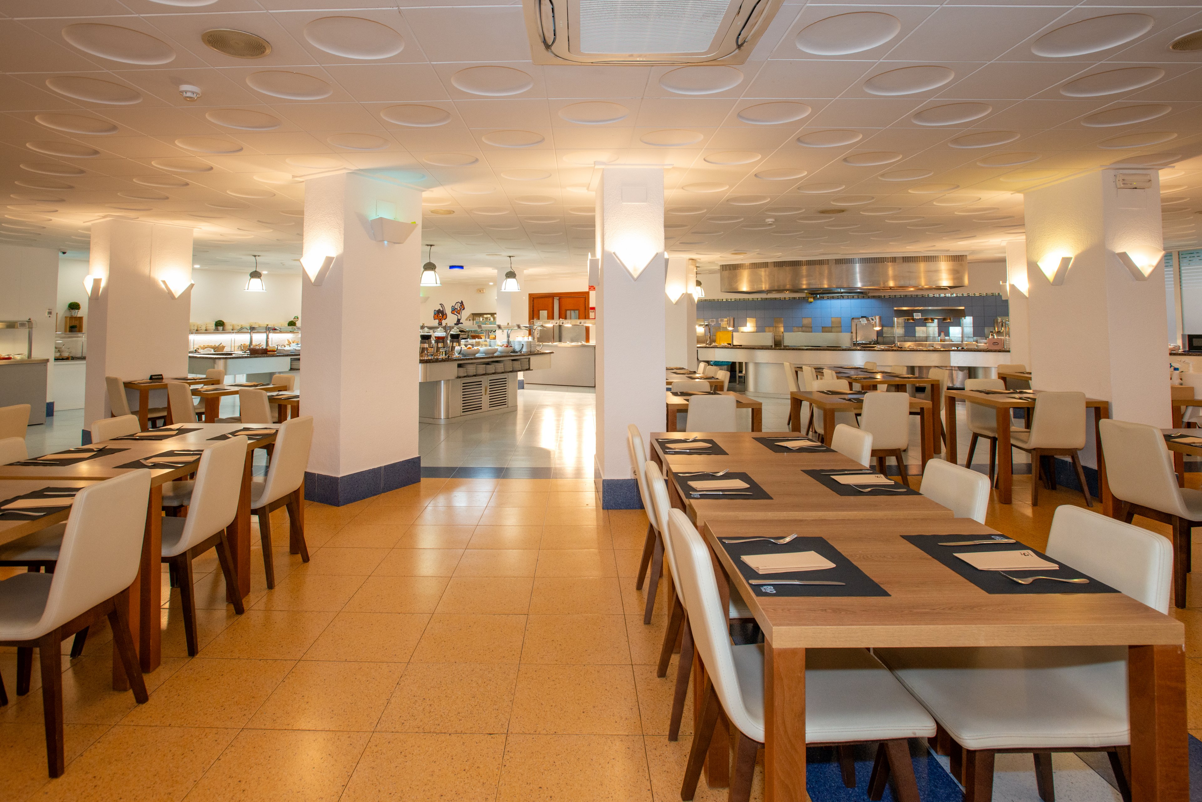 Spacious restaurant dining area with neatly arranged wooden tables and light-colored chairs. The room is empty of people, emphasizing the clean and orderly setup. Warm ceiling and pendant lights illuminate the space, leading the eye toward an open buffet or serving counter in the background. Contemporary decor and neutral tones create a welcoming, modern atmosphere suitable for hotel or cafeteria dining.