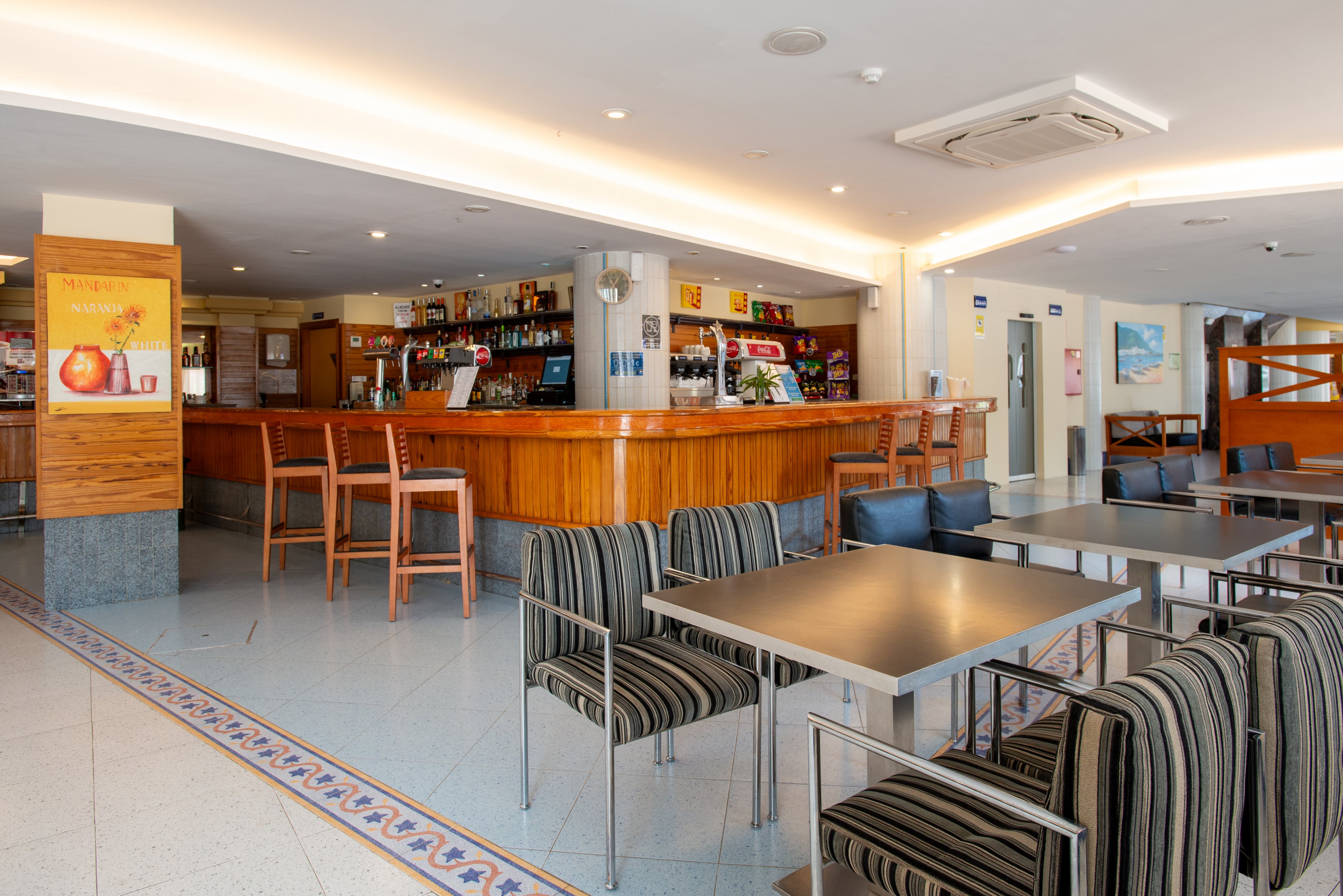 Spacious hotel bar interior featuring a long wooden counter and high stools. In the foreground, modern tables with striped upholstered chairs create a casual lounge area. The bar back displays various bottles, snacks, and a refrigerator, suggesting a relaxed refreshment space. Warm ceiling lighting and tiled floors contribute to a clean, welcoming atmosphere.