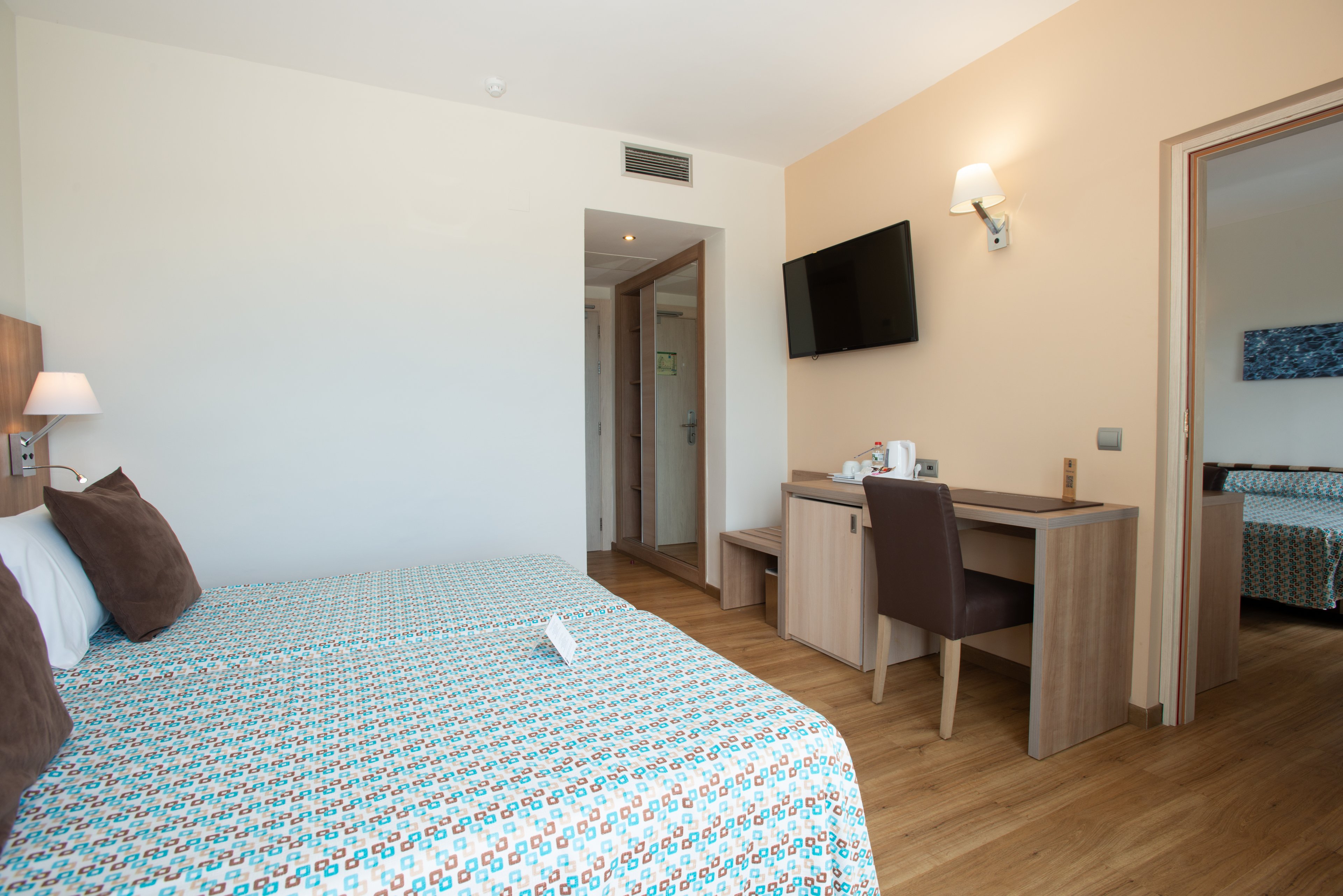 A bright modern hotel room features a neatly made double bed with patterned bedding. A wall-mounted flat-screen TV faces a wooden desk with a chair and a small hospitality tray. Light-colored walls and wood flooring create a clean, contemporary look. An open doorway reveals an adjoining bedroom with a similar bed and decor, suggesting a family or connecting room layout.