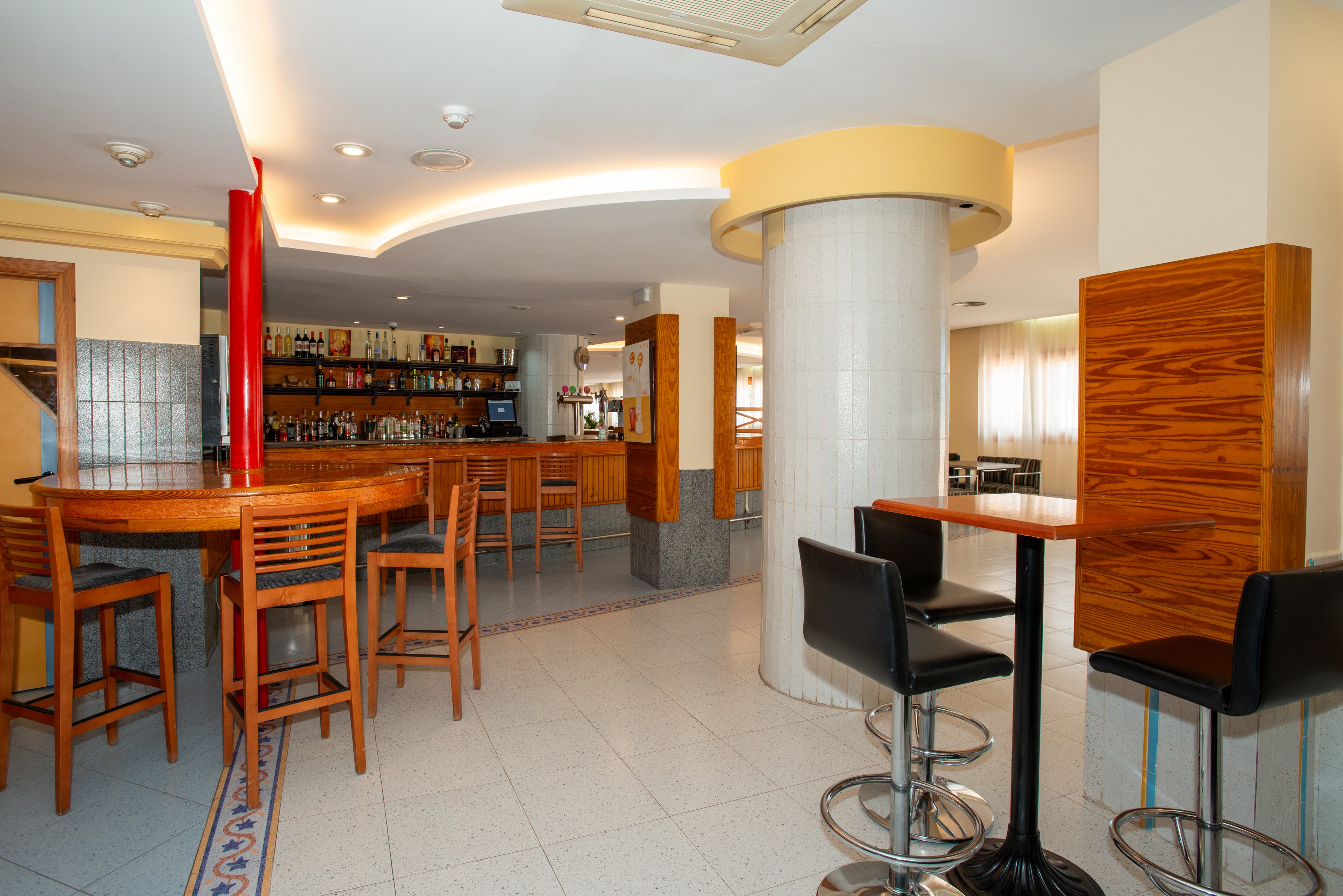 Contemporary bar interior featuring a long wooden counter and matching high tables. The space is empty, highlighting the clean tiled floor and curved ceiling lighting. A central column and light-colored walls divide the seating areas with both wooden stools and black bar chairs. Bottles and glassware are neatly arranged on shelves behind the bar, creating a tidy and inviting atmosphere.