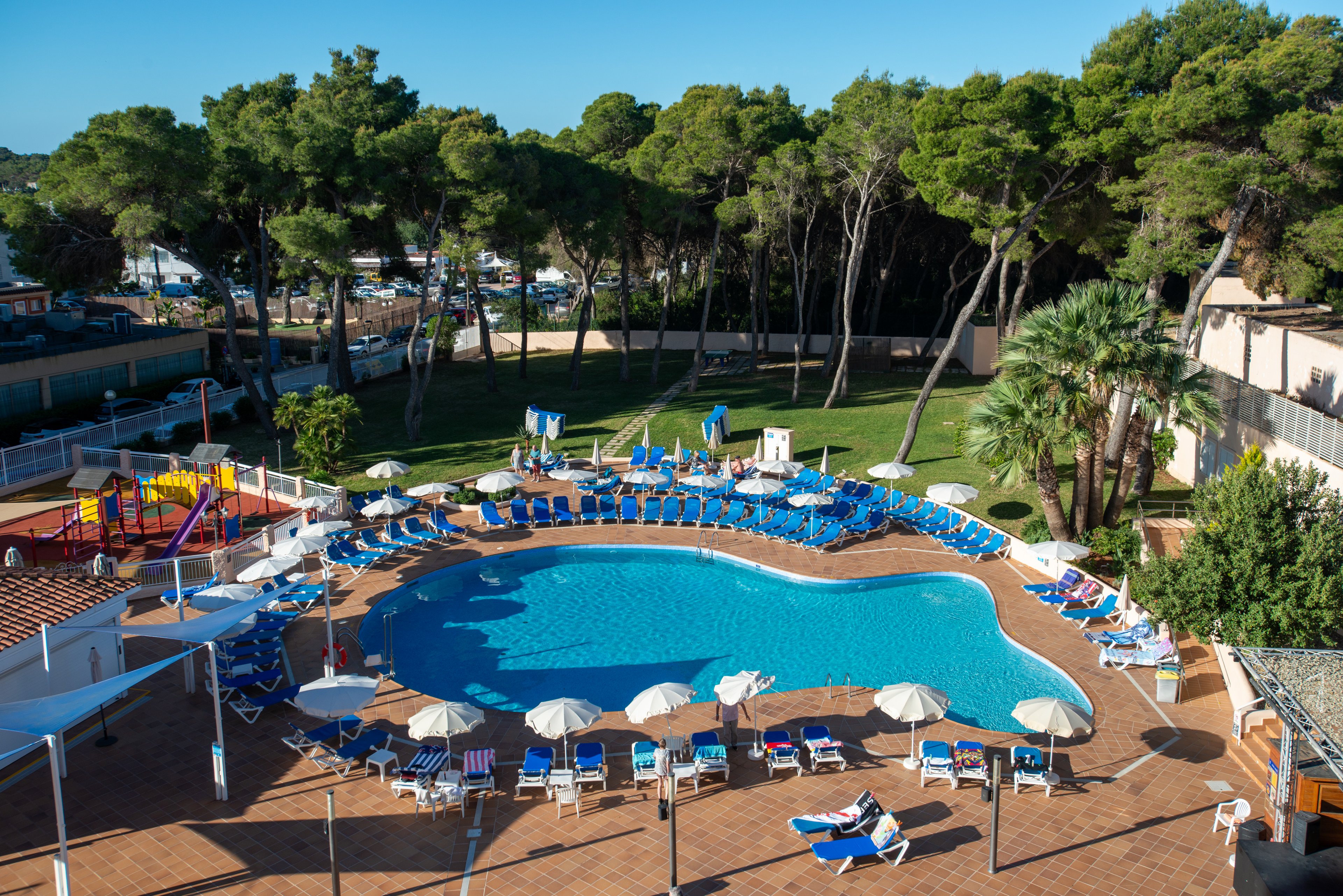 A large outdoor swimming pool is surrounded by rows of blue sun loungers and white umbrellas at a holiday resort. The scene is set on a spacious tiled terrace with a nearby children's playground featuring colorful slides and climbing structures. Tall green trees and a grassy lawn create a natural backdrop around the pool area. The bright daylight and clear blue water give the setting a relaxed, summery vacation atmosphere.