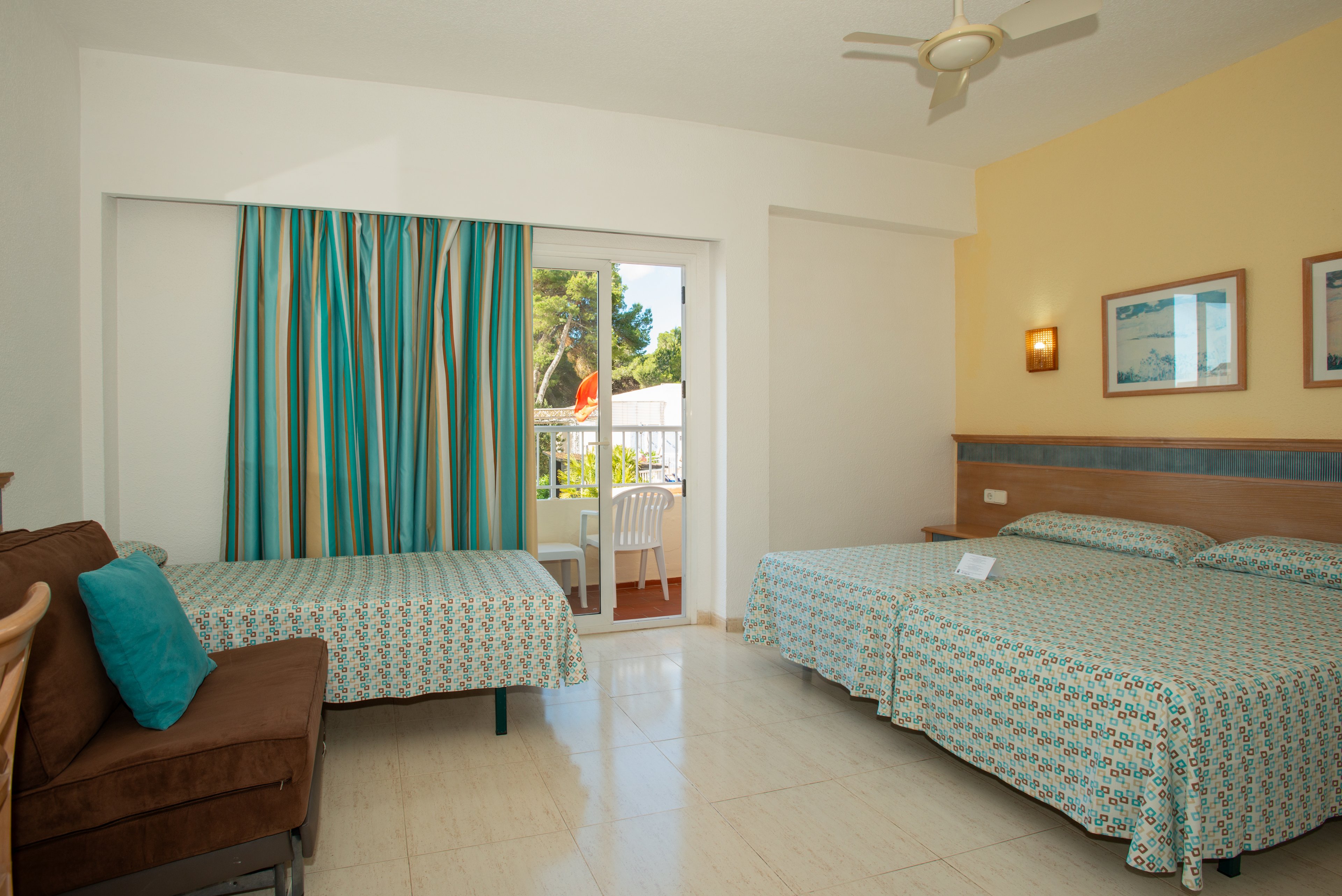A bright hotel room features two twin beds with matching patterned bedspreads and a small sofa with teal cushions. The space opens onto a balcony through a sliding glass door, where white patio chairs and a table are visible. Striped curtains frame the doorway, and a ceiling fan and wall-mounted light add comfort and ambiance. The room has light-colored tile flooring and simple framed artwork on the walls, creating a clean and inviting atmosphere.