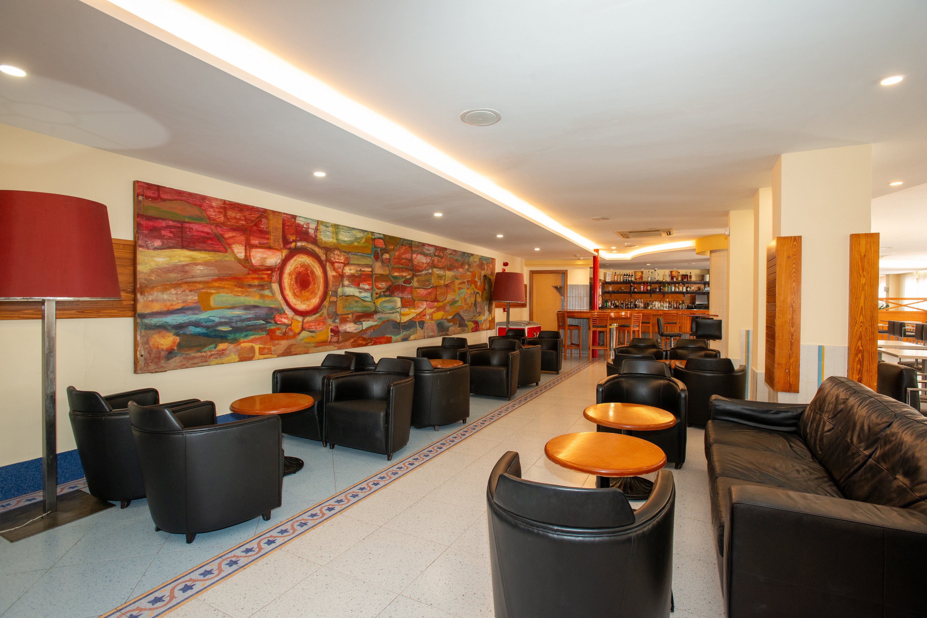 Spacious hotel lounge area featuring black leather armchairs and small round wooden tables. A long, colorful abstract mural with a sun-like motif dominates one wall, adding warmth and visual interest. Soft ceiling lighting and large red floor lamps create a cozy, inviting atmosphere. In the background, a bar counter with bottles and stools suggests a social gathering space.