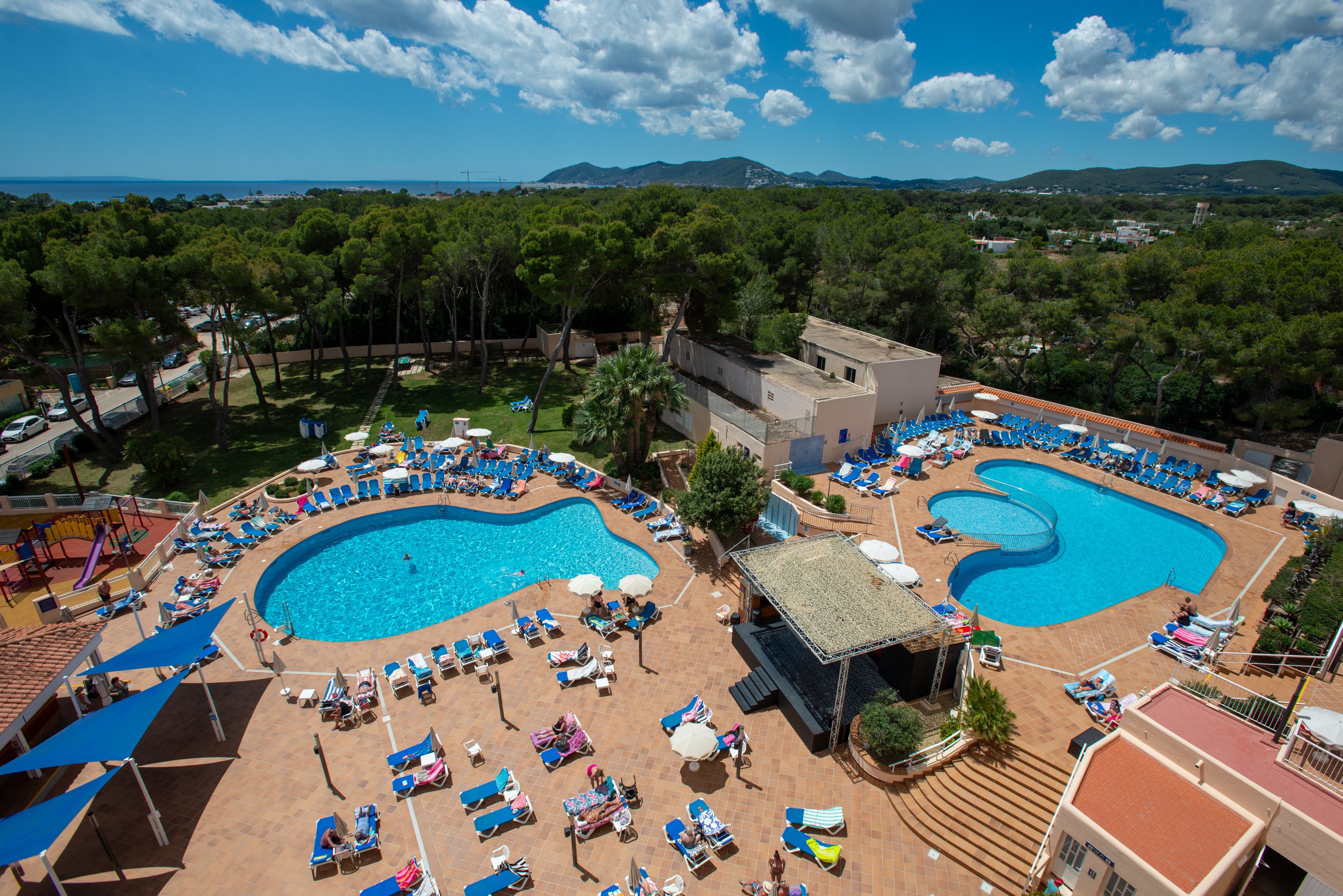 A large holiday resort features two outdoor swimming pools surrounded by sun loungers and umbrellas. Guests relax on deck chairs, sunbathe, and swim in the bright blue water under a sunny sky. The pool terrace is bordered by tall green trees, with distant hills and the sea visible on the horizon. The scene conveys a lively, family-friendly vacation atmosphere in a warm coastal destination.