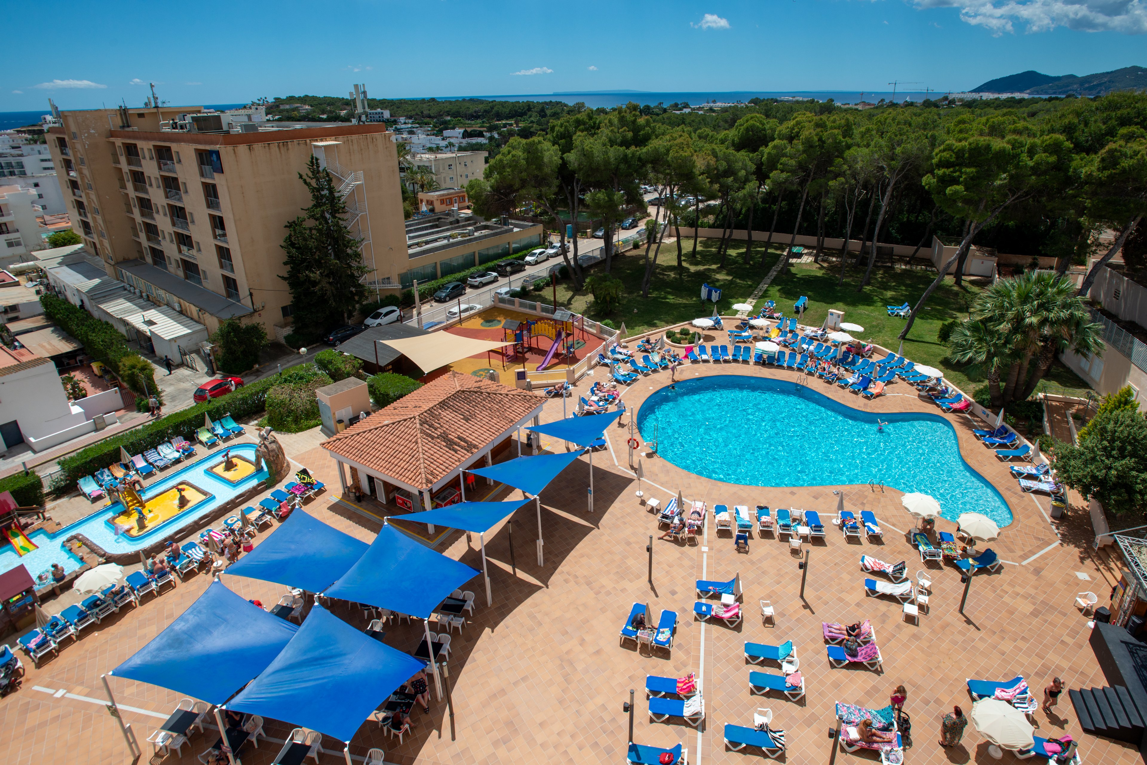 An aerial view shows a lively resort swimming pool surrounded by numerous sun loungers and parasols. Families and holidaymakers relax and sunbathe around the curved blue pool while children enjoy a nearby splash play area. Blue shade canopies cover part of the terrace, and colorful buildings and trees frame the scene. In the distance, a coastal town and the sea are visible under a bright, clear sky.