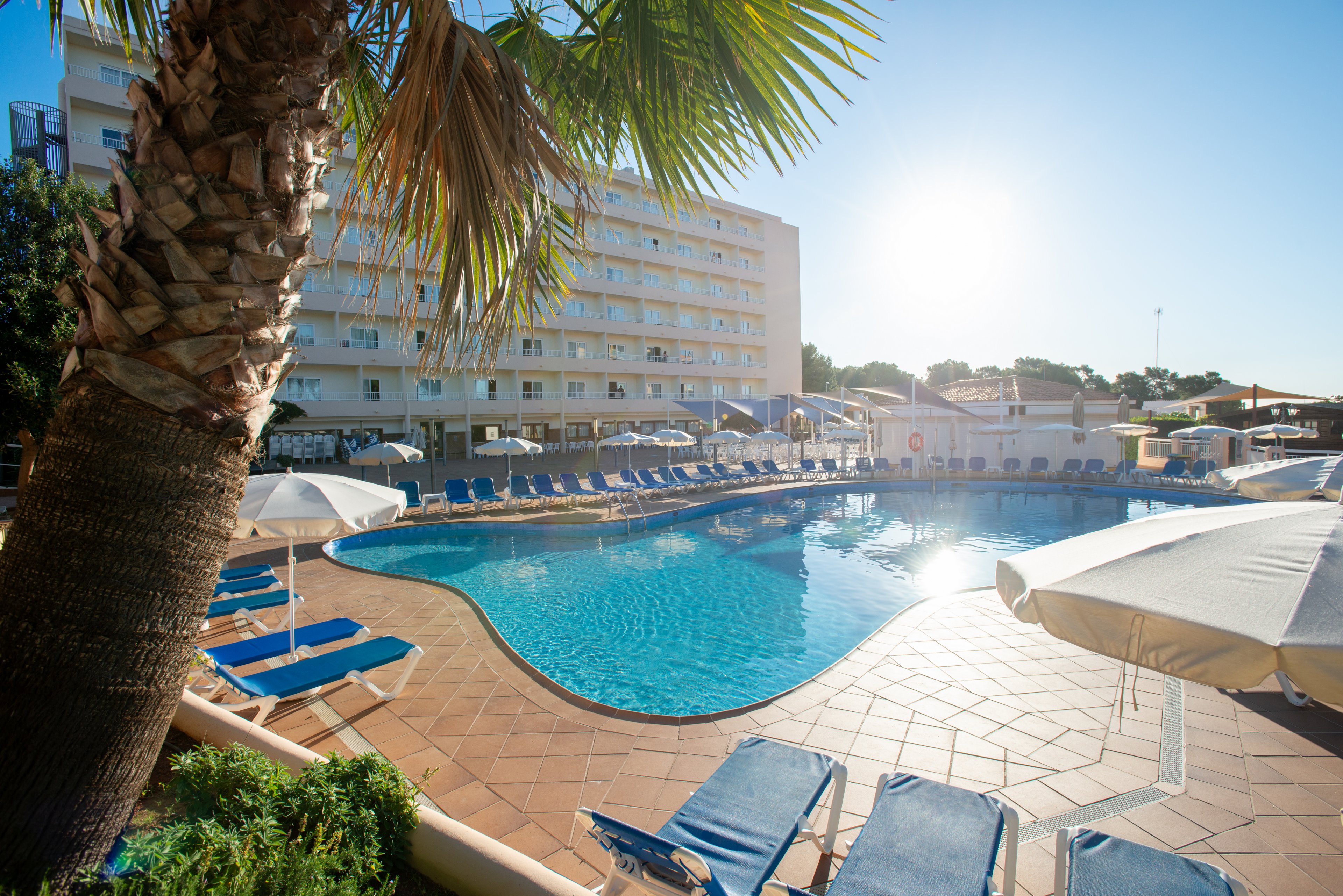 A large outdoor swimming pool curves through the center of a resort hotel courtyard. Blue sun loungers and white umbrellas surround the pool on a tiled terrace. A multi-story hotel building rises in the background under a clear blue sky. Bright sunlight creates strong reflections on the water, suggesting a warm, relaxing vacation atmosphere.
