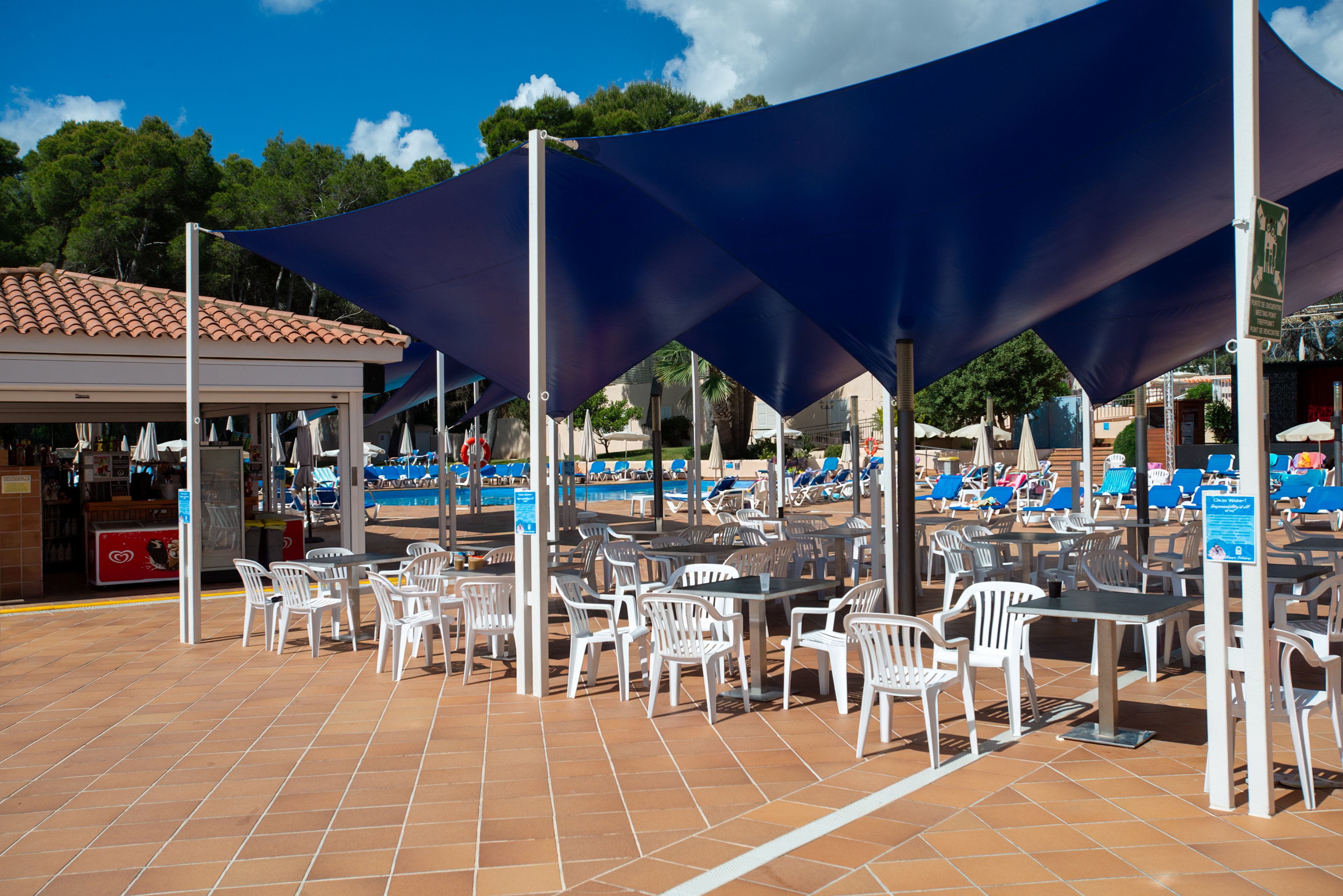 An outdoor resort terrace features rows of white plastic tables and chairs beneath large blue shade sails. The seating area is adjacent to a swimming pool filled with sun loungers and guests enjoying the sunny weather. Terracotta tiles cover the ground, and a small bar or snack kiosk stands to the side. Lush green trees and a bright blue sky with scattered clouds frame the relaxed holiday setting.