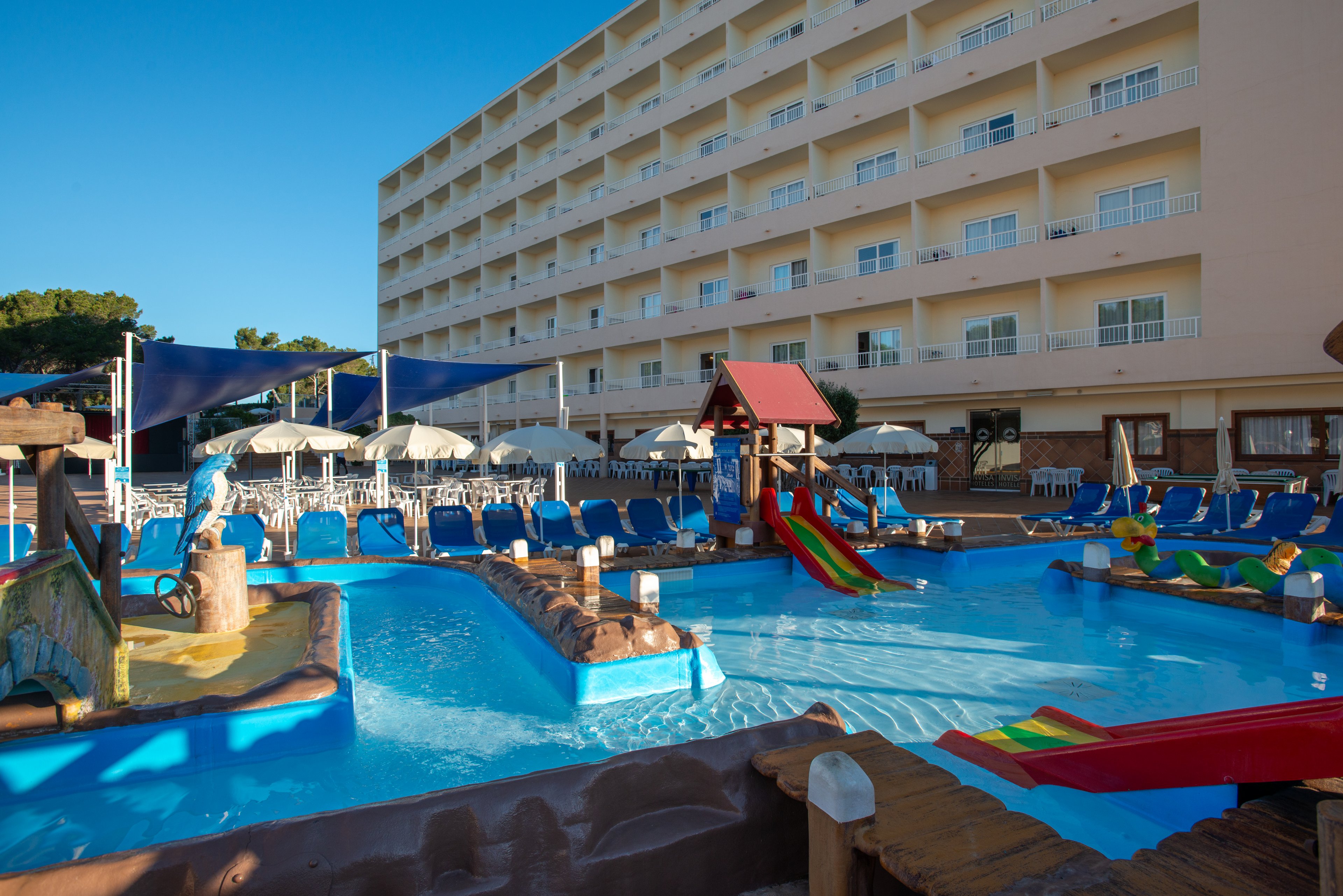 An outdoor children's splash pool sits in front of a multi-story hotel building with balconies. The pool features colorful slides, shallow water, and playful structures designed for kids. Rows of blue sun loungers and white umbrellas line the poolside under shade canopies. The scene is brightly lit with clear blue sky, suggesting a warm vacation setting.