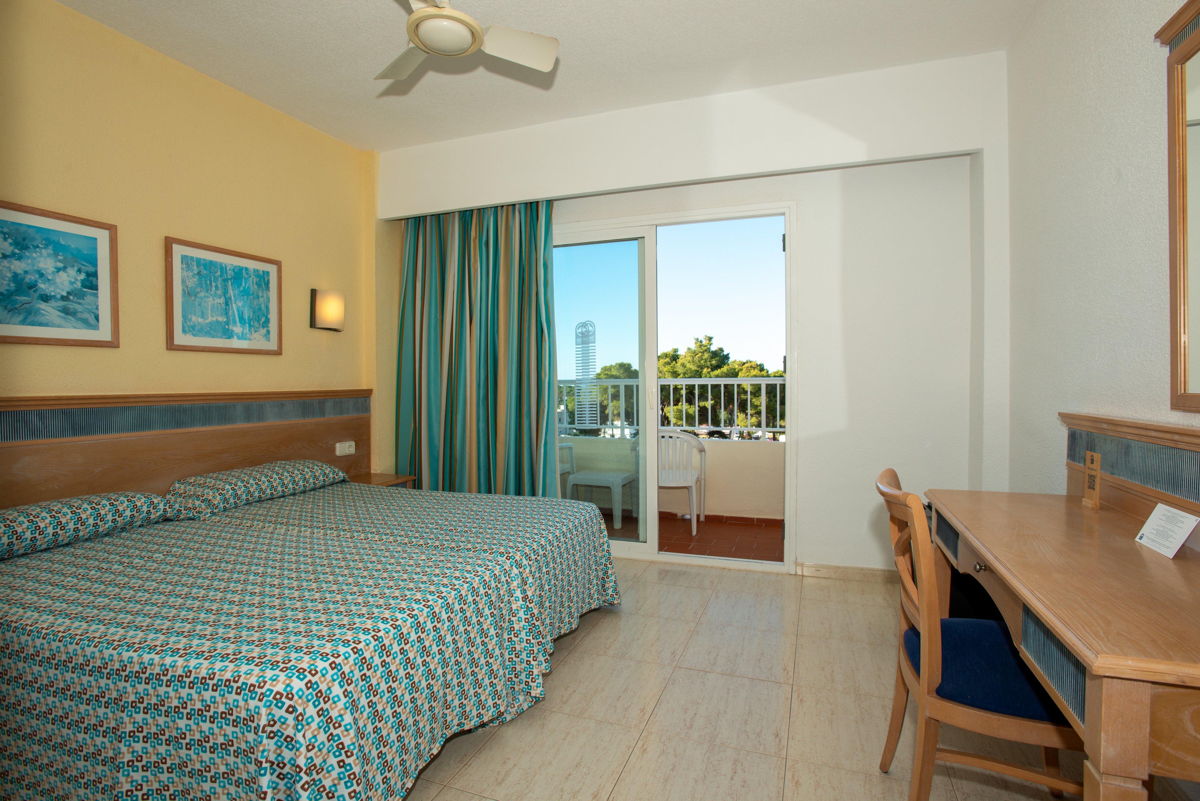 A tidy hotel room features two beds pushed together with patterned bedspreads. A large sliding glass door opens to a balcony with outdoor chairs and a view of trees and distant buildings. The interior includes a wooden desk with a chair, wall art, and a ceiling fan. Natural light and blue-green accents create a fresh, inviting atmosphere.