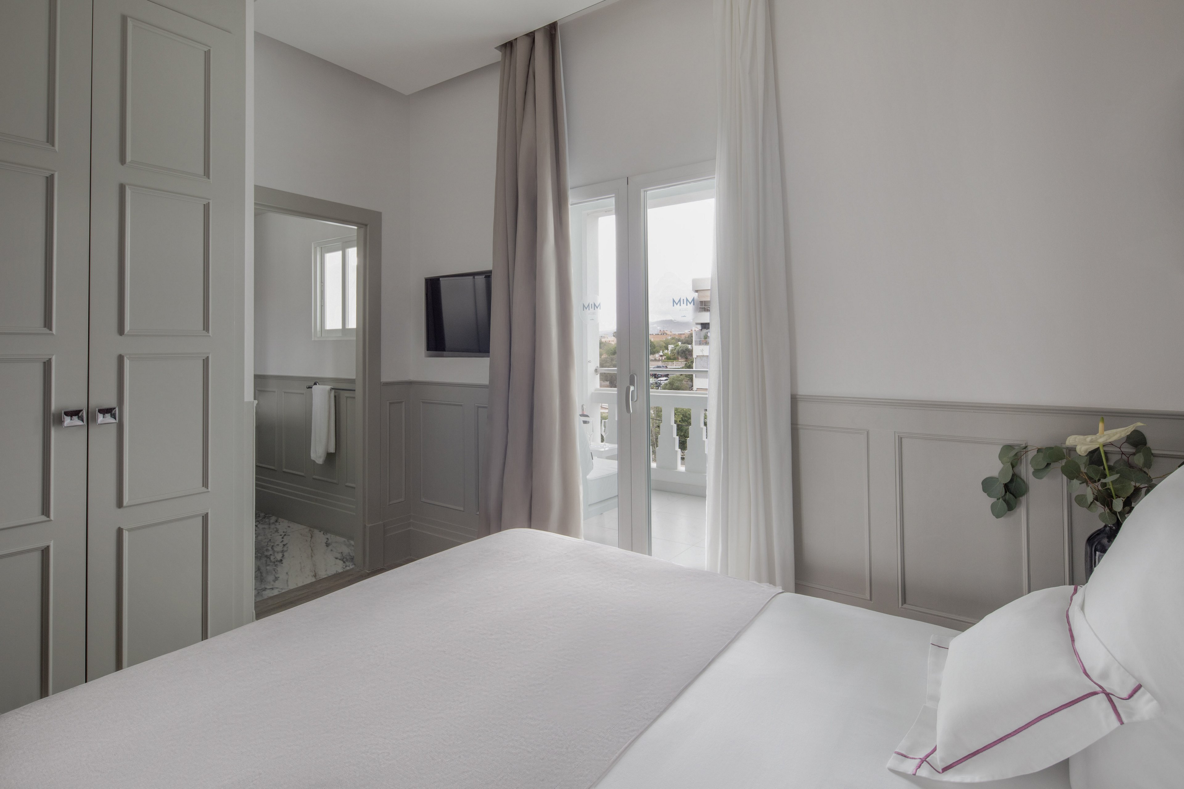 A bright and modern bedroom featuring a neatly made bed with white linens and subtle accents. The room includes a wall-mounted TV, a wardrobe, and a balcony with sheer curtains allowing natural light to flow in. Neutral tones and clean lines create a serene and minimalist aesthetic.