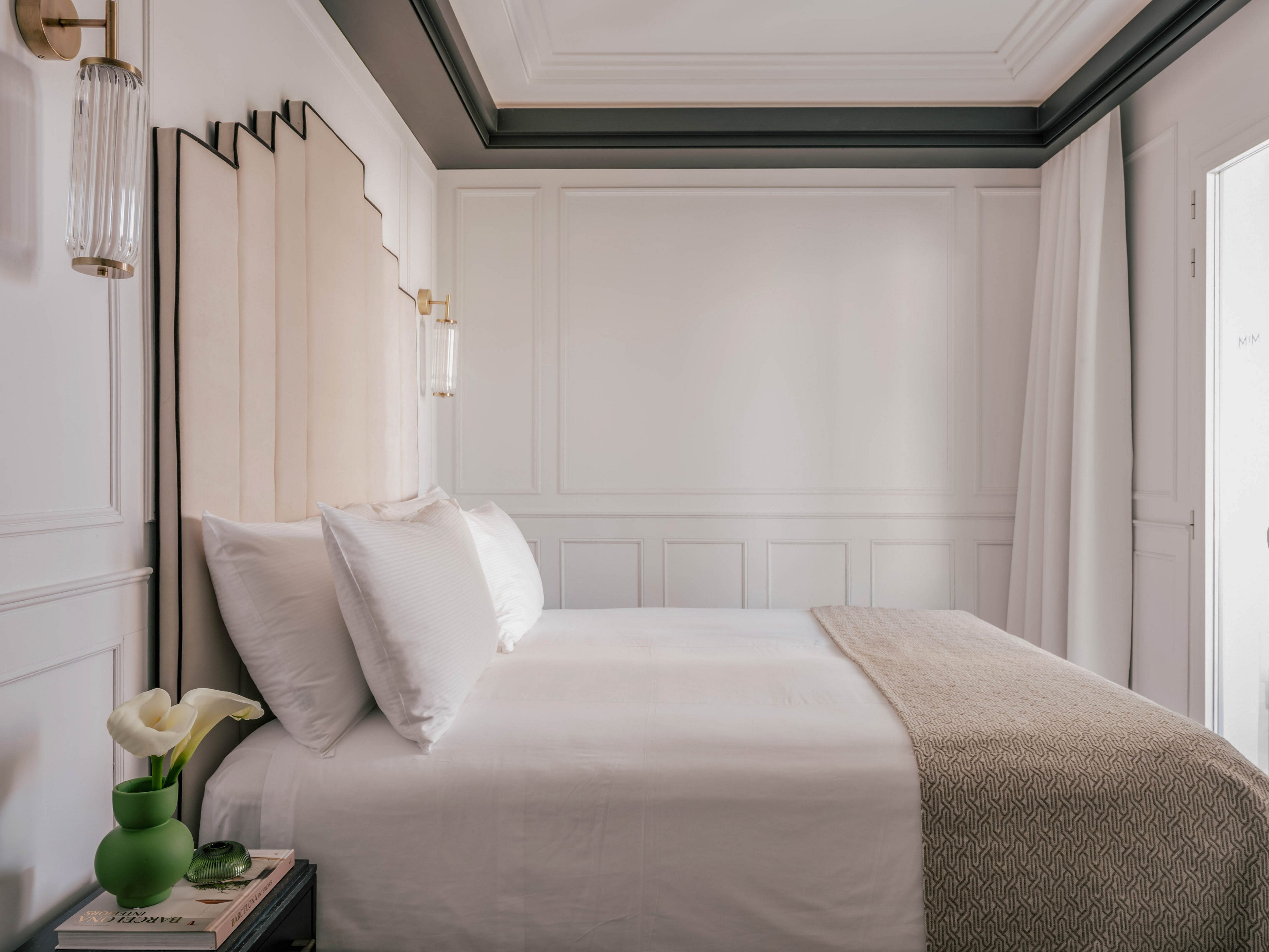 A neatly made bed with crisp white linens is positioned against a paneled wall in a minimalist bedroom. The room features soft neutral tones, including a beige headboard and a textured beige throw blanket. Elegant wall sconces provide warm lighting, and a small bedside table holds a green vase with white flowers and a book. Natural light enters from a doorway or window on the right, enhancing the calm and serene atmosphere.