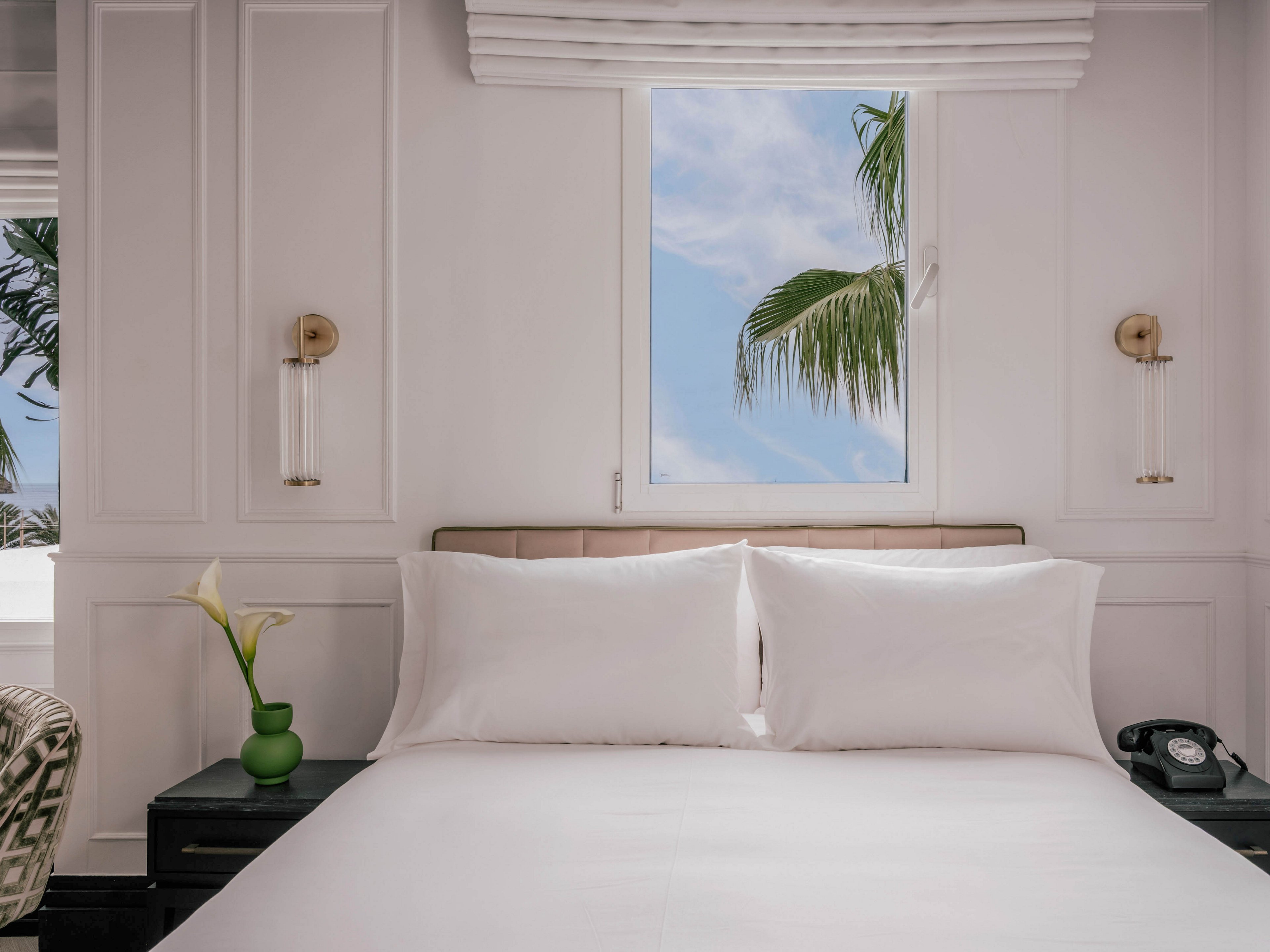 A neatly made bed with crisp white linens is centered in a bright, modern bedroom. The room features white paneled walls, minimal decor, and a window framing a clear sky with palm tree leaves outside. Matching bedside tables hold a green vase with calla lilies and a classic black telephone, adding subtle color accents. Soft natural light enhances the serene, tropical atmosphere of the space.