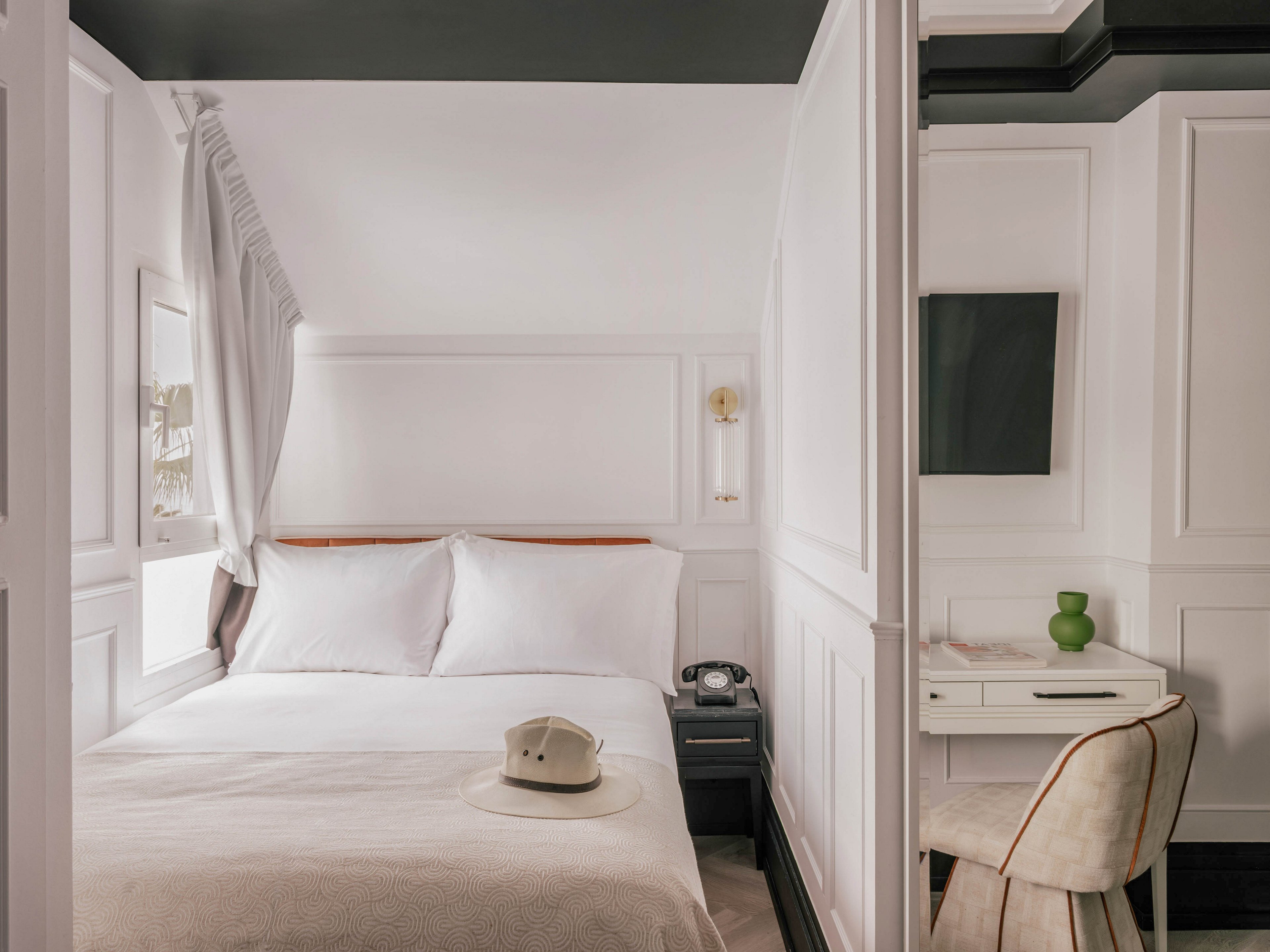 A compact hotel room features a neatly made double bed with white linens and a light blanket. A small bedside table holds a vintage-style telephone, while a hat rests on the bed. To the right, a built-in desk with a chair, green vase, and wall-mounted television creates a functional workspace. The room’s white paneled walls and soft neutral tones contribute to a calm, minimalist atmosphere.