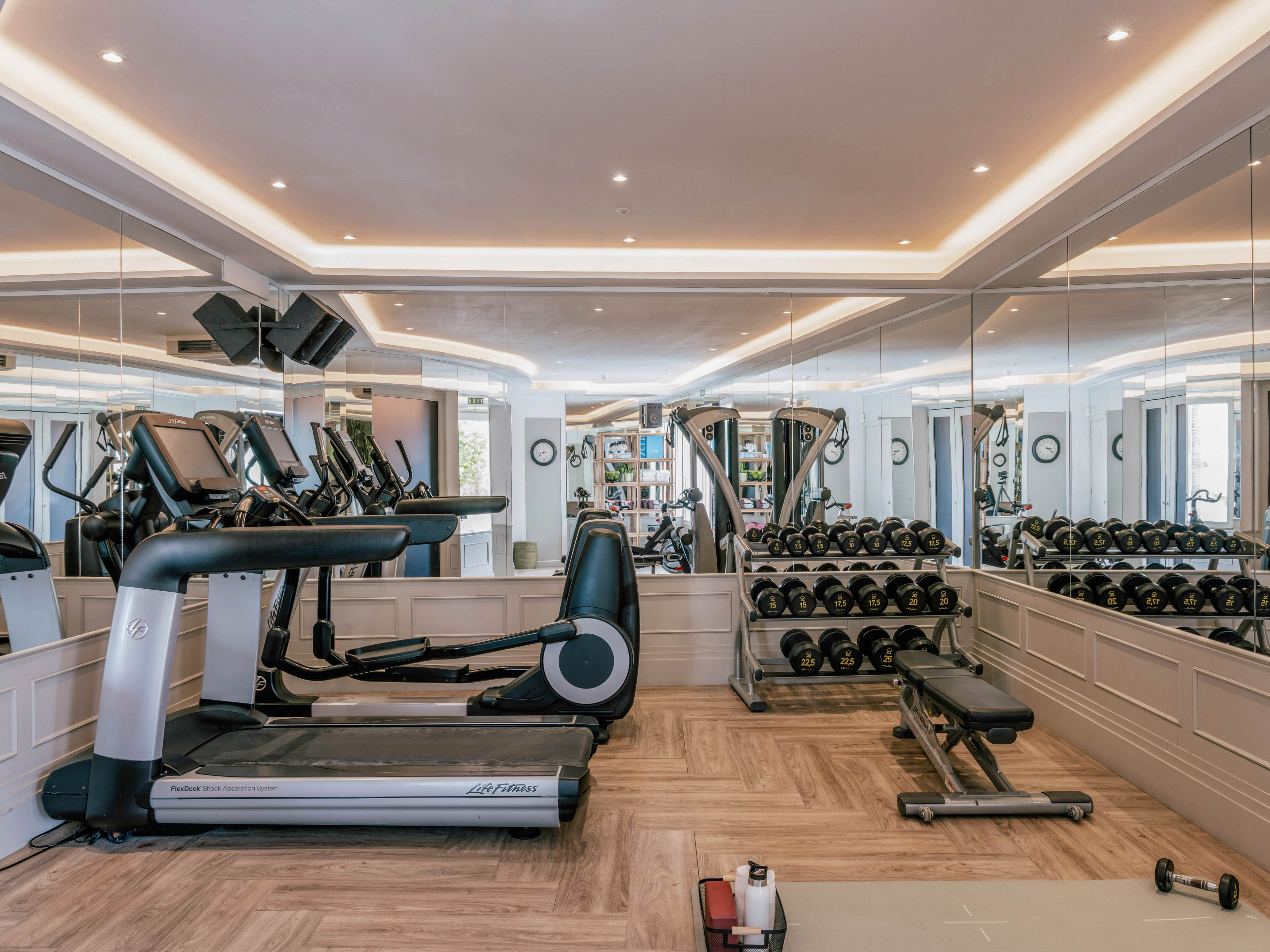 A sleek home gym features a large treadmill and a bench positioned in front of mirrored walls. Multiple racks of dumbbells line the right side, reflecting in the mirrors to create a spacious feel. Warm recessed ceiling lights and light wood flooring give the room a clean, contemporary look. The space appears organized and ready for personal fitness workouts.