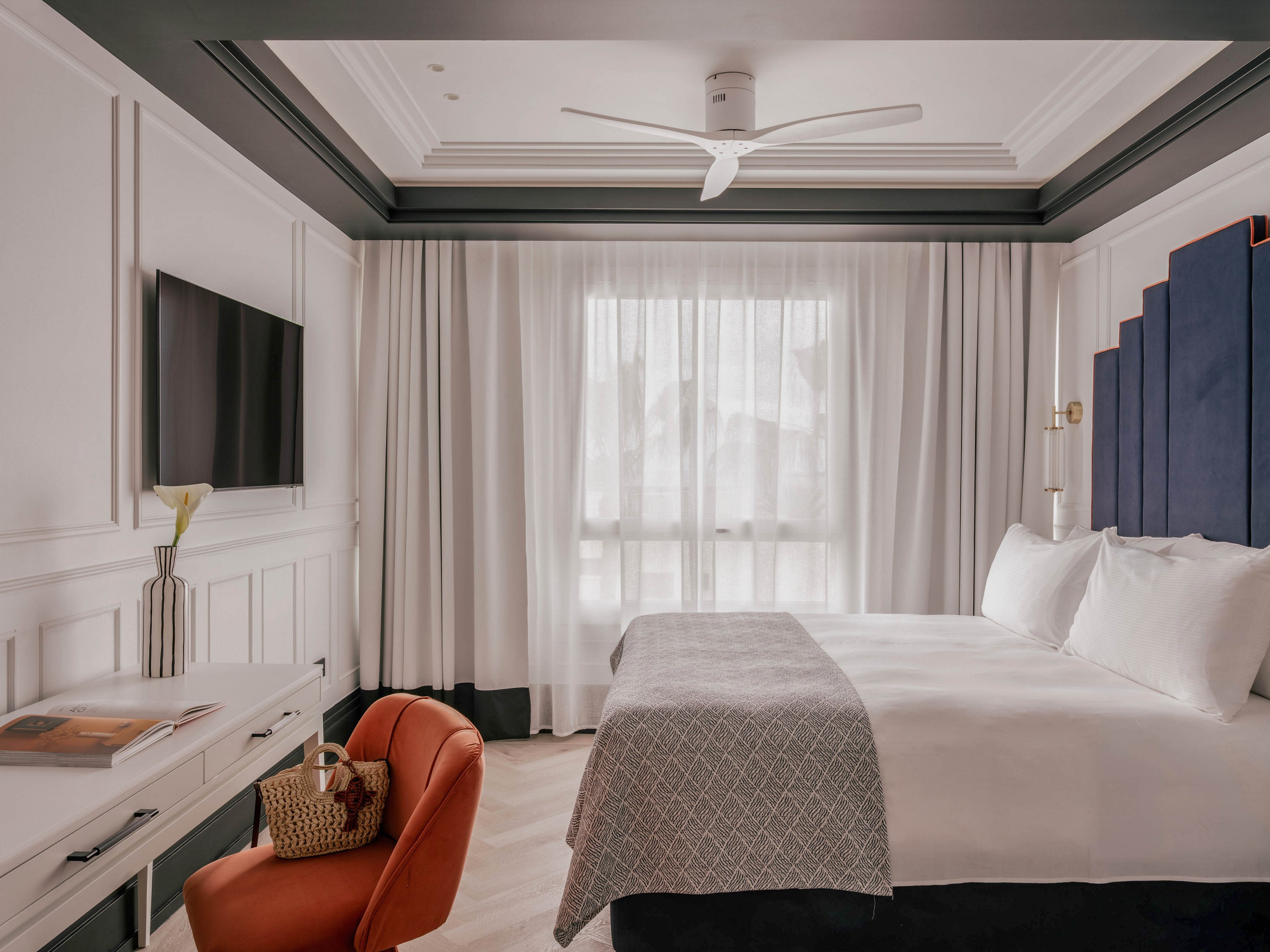 A contemporary hotel bedroom features a neatly made bed with white linens and a textured gray blanket. Soft daylight filters through sheer curtains covering a large window, brightening the neutral-toned room. A wall-mounted television, ceiling fan, and built-in desk with a book and woven bag add functional details. An orange accent chair and dark headboard panels provide contrast against the light walls and flooring, creating a calm, inviting atmosphere.