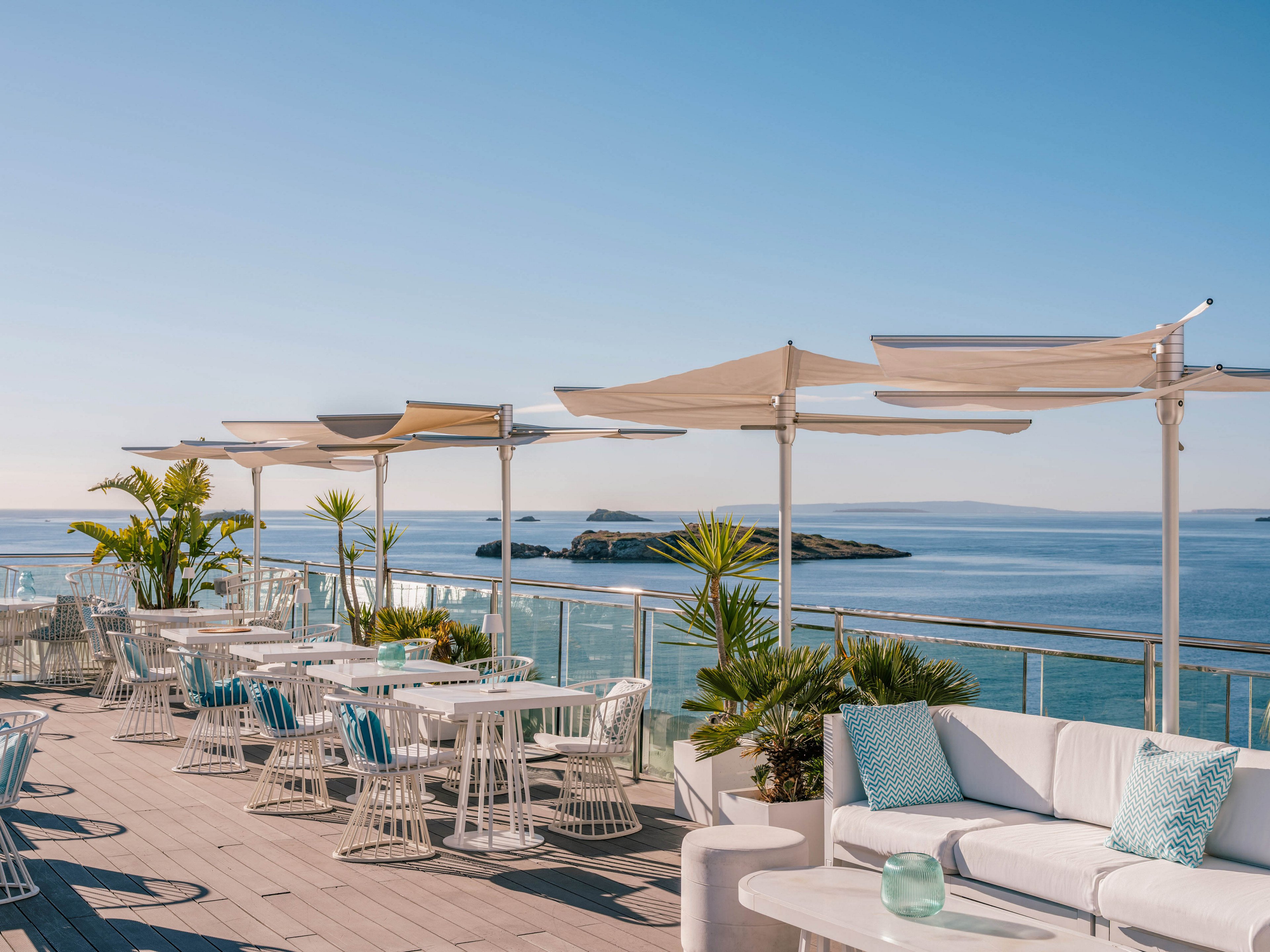 An open-air rooftop terrace overlooks a calm blue sea under a clear sky. White tables, chairs, and cushioned sofas are arranged neatly along the deck, accented with light blue pillows. Modern shade canopies provide partial cover, while glass railings frame the ocean view and distant islands. Potted tropical plants add greenery and a relaxed, resort-like atmosphere.