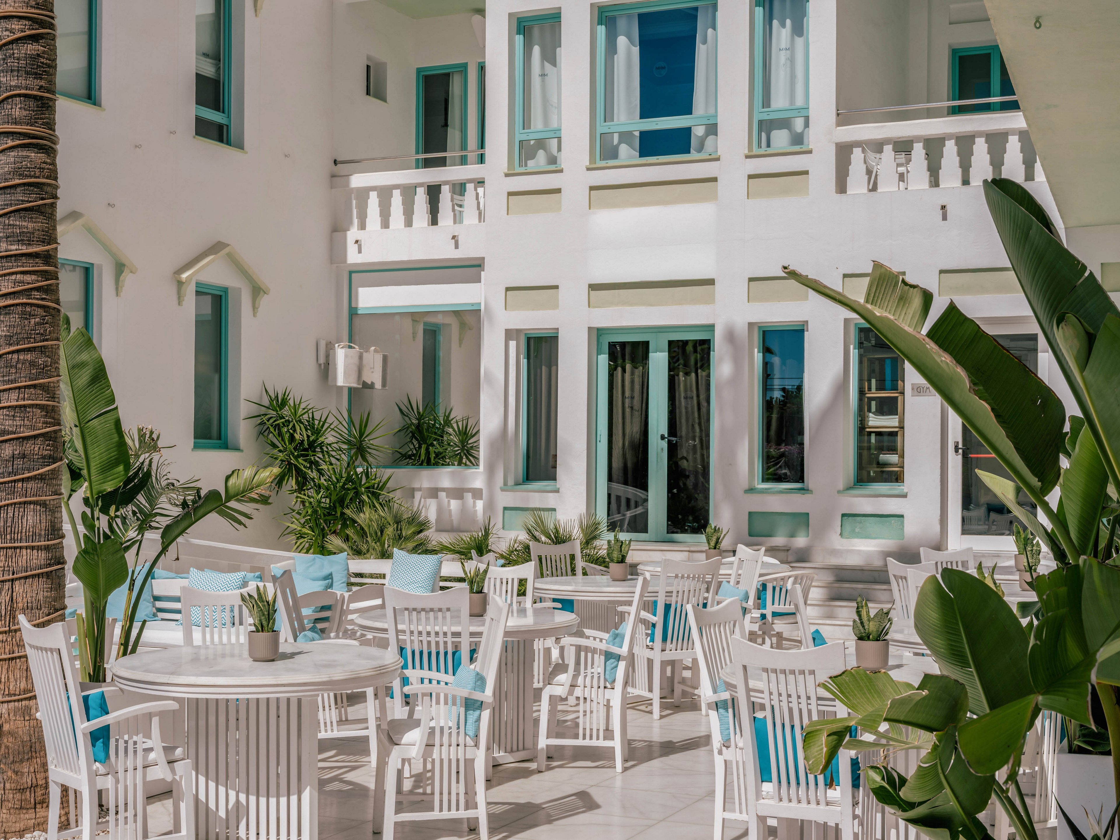 An outdoor courtyard cafe features white wooden tables and chairs arranged neatly under bright daylight. The setting is framed by a white building with pastel blue window frames and balconies, giving a coastal resort feel. Potted plants and tropical greenery add natural accents around the seating area. The overall atmosphere is calm, inviting, and ideal for relaxed dining or socializing.
